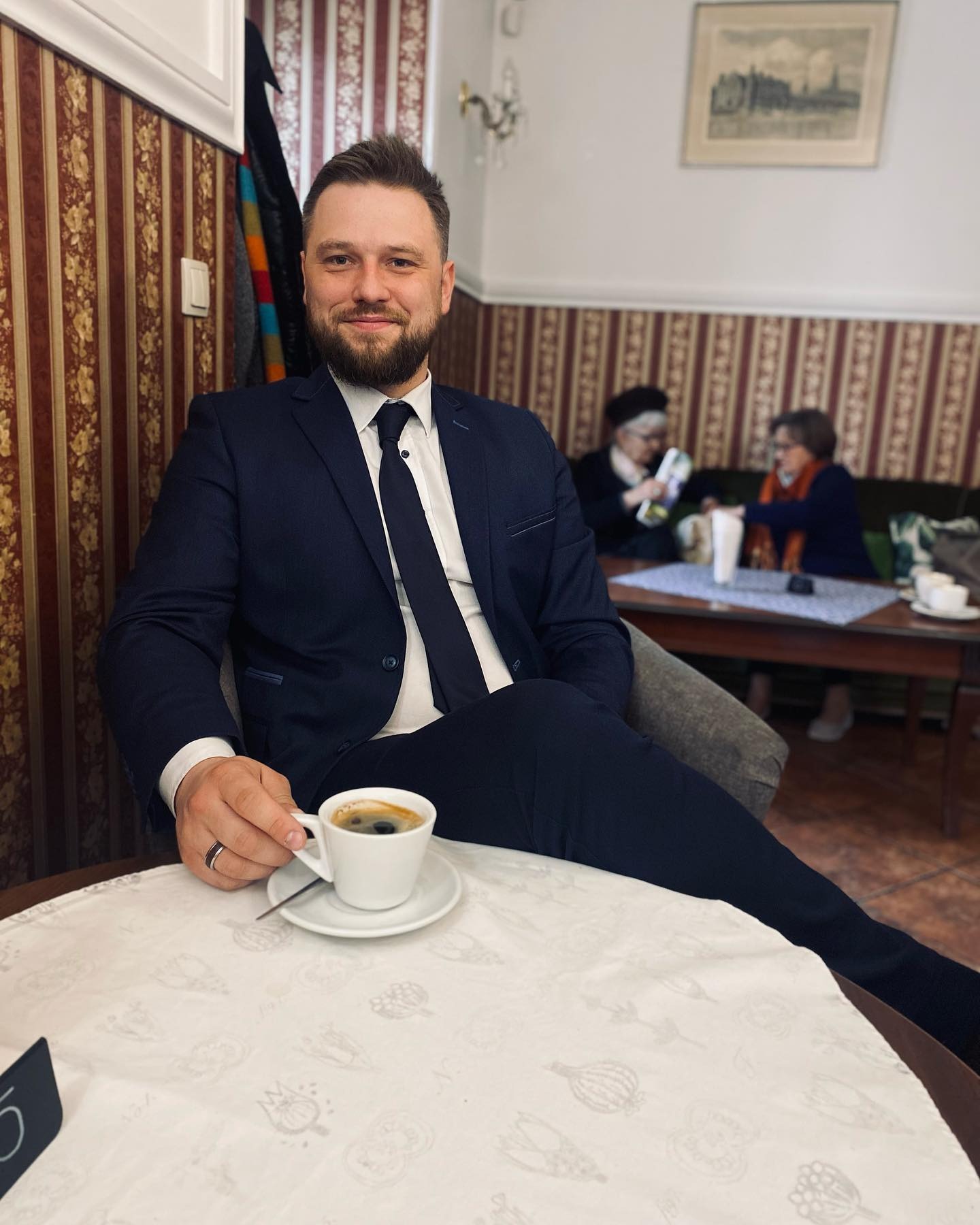 Uśmiechnięty mężczyzna w garniturze pije kawę w kawiarni z dwoma starszymi paniami w tle, siedząc przy okrągłym stoliku z białym obrusem w kwiatowe wzory.
