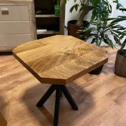 Ośmiokątny stolik kawowy z blatem z jasnego drewna o wyraźnym usłojeniu i czarnymi, metalowymi nogami w kształcie trójnogu, na tle drewnianej podłogi, komody i roślin doniczkowych.