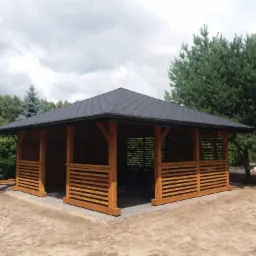 Nowoczesna, drewniana konstrukcja z czarnym dachem bitumicznym, otoczona zielenią, z widocznymi elementami wykończeniowymi z jasnego drewna i szarą kostką brukową