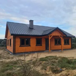 Nowo wybudowany, parterowy dom z drewna o pomarańczowej barwie, z czarnymi ramami okiennymi i metalowym dachem w kolorze grafitowym, widoczny komin i zadaszone wejście, surowy teren wokół budynku.