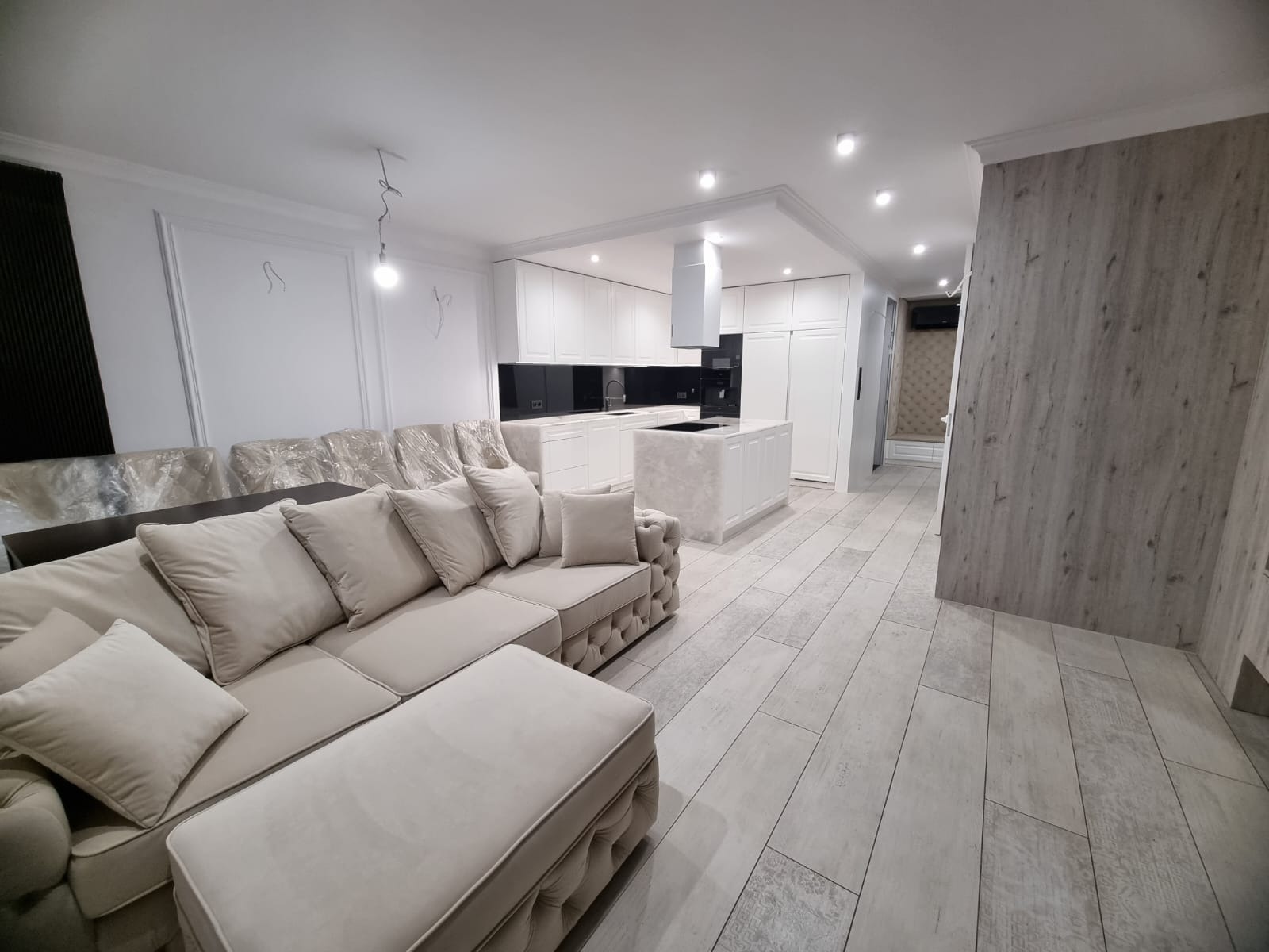 Jasne wnętrze z sofą obitą tkaniną w stylu Chesterfield, częściowo owiniętą folią ochronną, i białą zabudową kuchenną z wyspą, podłoga wyłożona panelami imitującymi drewno.