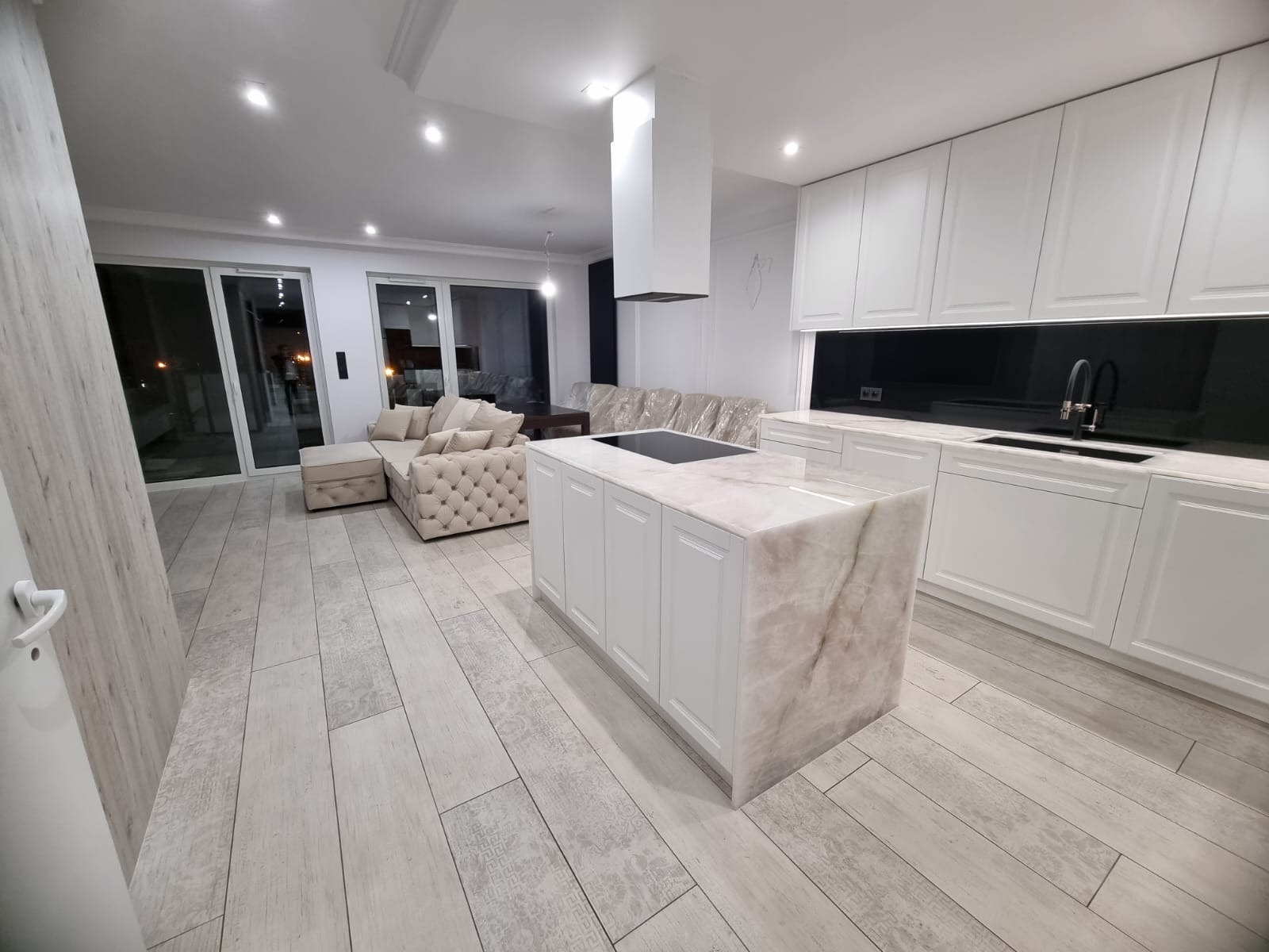 Nowoczesne wnętrze w Lesznie z białą kuchnią z wyspą o marmurowym blacie, widok na salon z beżową sofą i duże okna przesuwne.