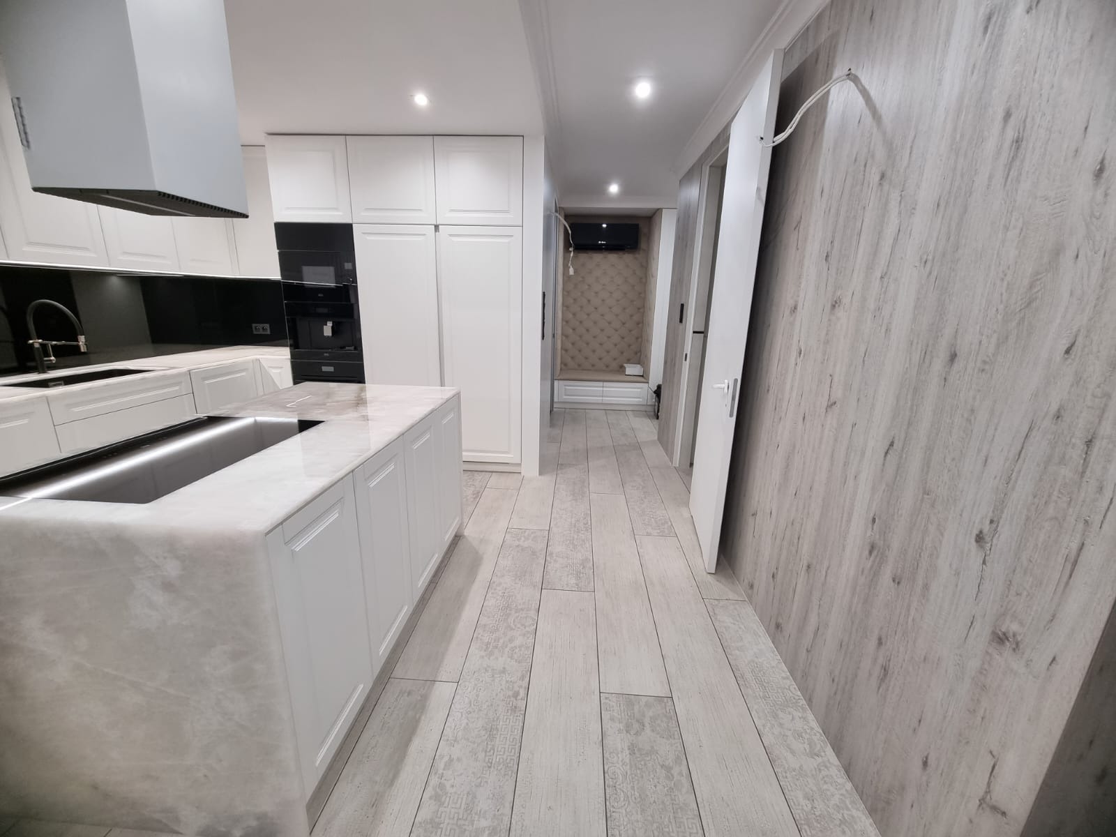 Nowoczesna kuchnia z białą zabudową, wyspą z marmurowym blatem i podświetlanym zlewem, połączona z korytarzem wykończonym panelami ściennymi imitującymi drewno.