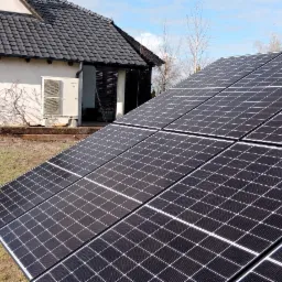 Czarny panel słoneczny na trawniku przed domem z pompą ciepła na ścianie, w tle błękitne niebo.