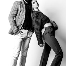 Kamil Tust - Czarno-białe zdjęcie pary w modnych ubraniach: mężczyzna w kraciastej marynarce i jasnych spodniach, kobieta w krótkiej kraciastej bluzce i spodniach z wysokim stanem, oboje opierają się o białą...