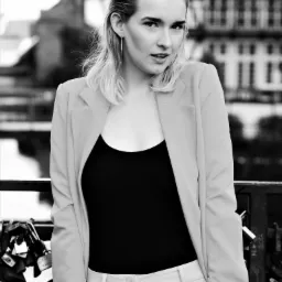 Czarno-białe portretowe ujęcie kobiety w marynarce i topie na tle miejskiego krajobrazu z mostem i kłódkami.