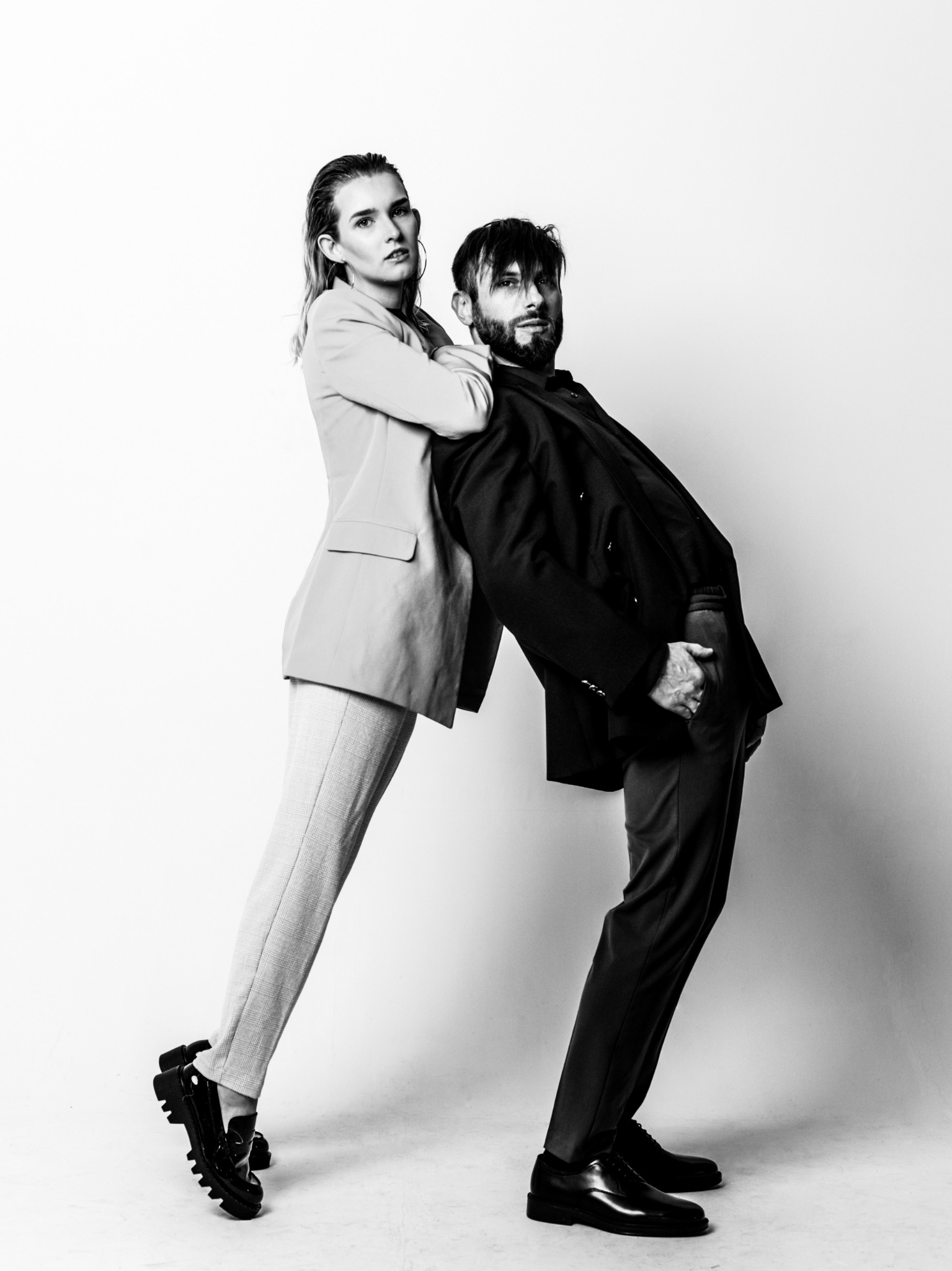 Czarno-białe zdjęcie dwojga modeli opartych o siebie plecami, ona w jasnym garniturze i masywnych butach, on w ciemnym garniturze i lakierkach, na białym tle.