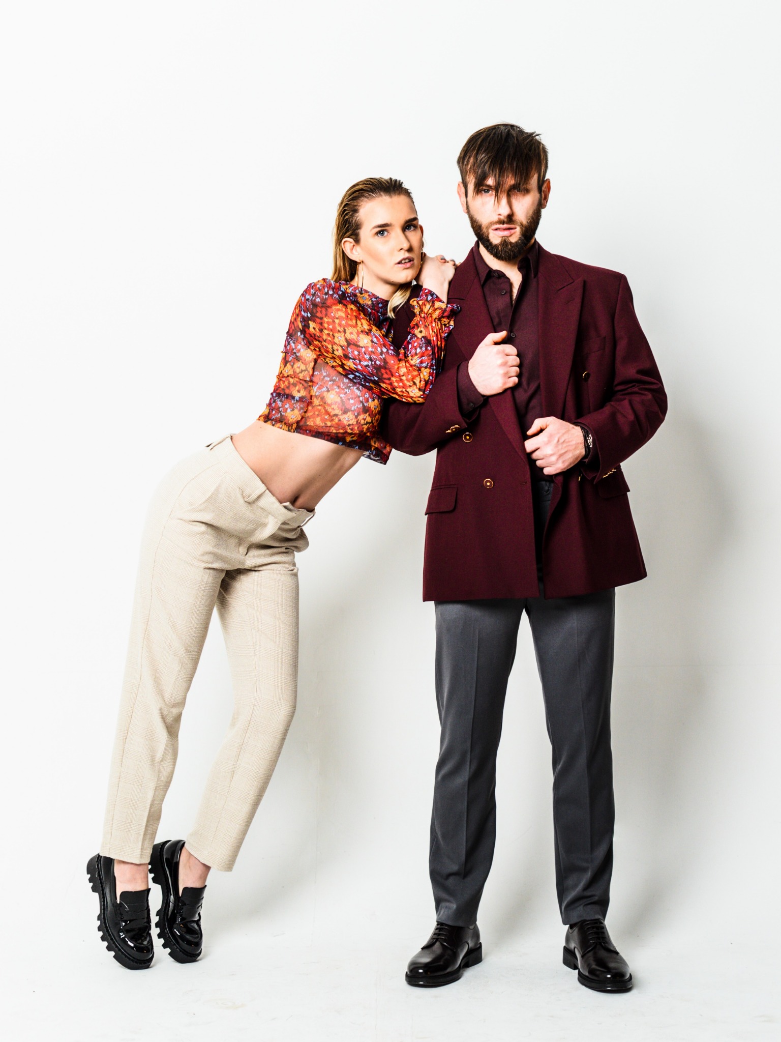 Para w studyjnej stylizacji: kobieta w transparentnej bluzce i spodniach w kratę, oparta o mężczyznę w bordowej marynarce i szarych spodniach.