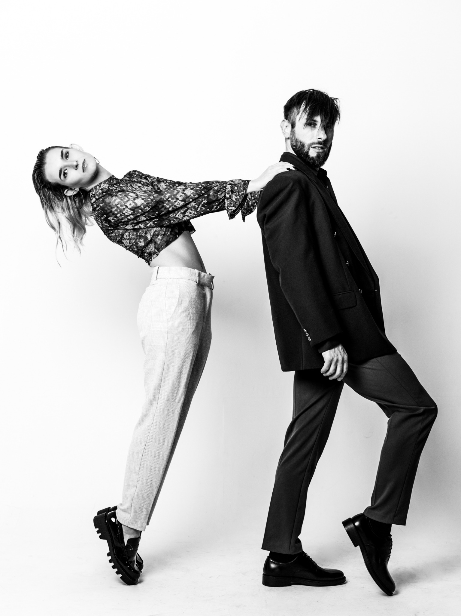 Czarno-białe zdjęcie studyjne pary w dynamicznej pozie: kobieta opiera się o ramię mężczyzny, oboje w eleganckich strojach na białym tle.