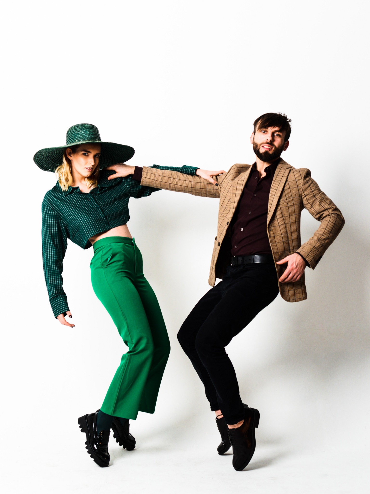 Para w modnych pozach na białym tle: kobieta w zielonym komplecie z dużym kapeluszem i mężczyzna w brązowej marynarce w kratę, oboje w eleganckich butach.
