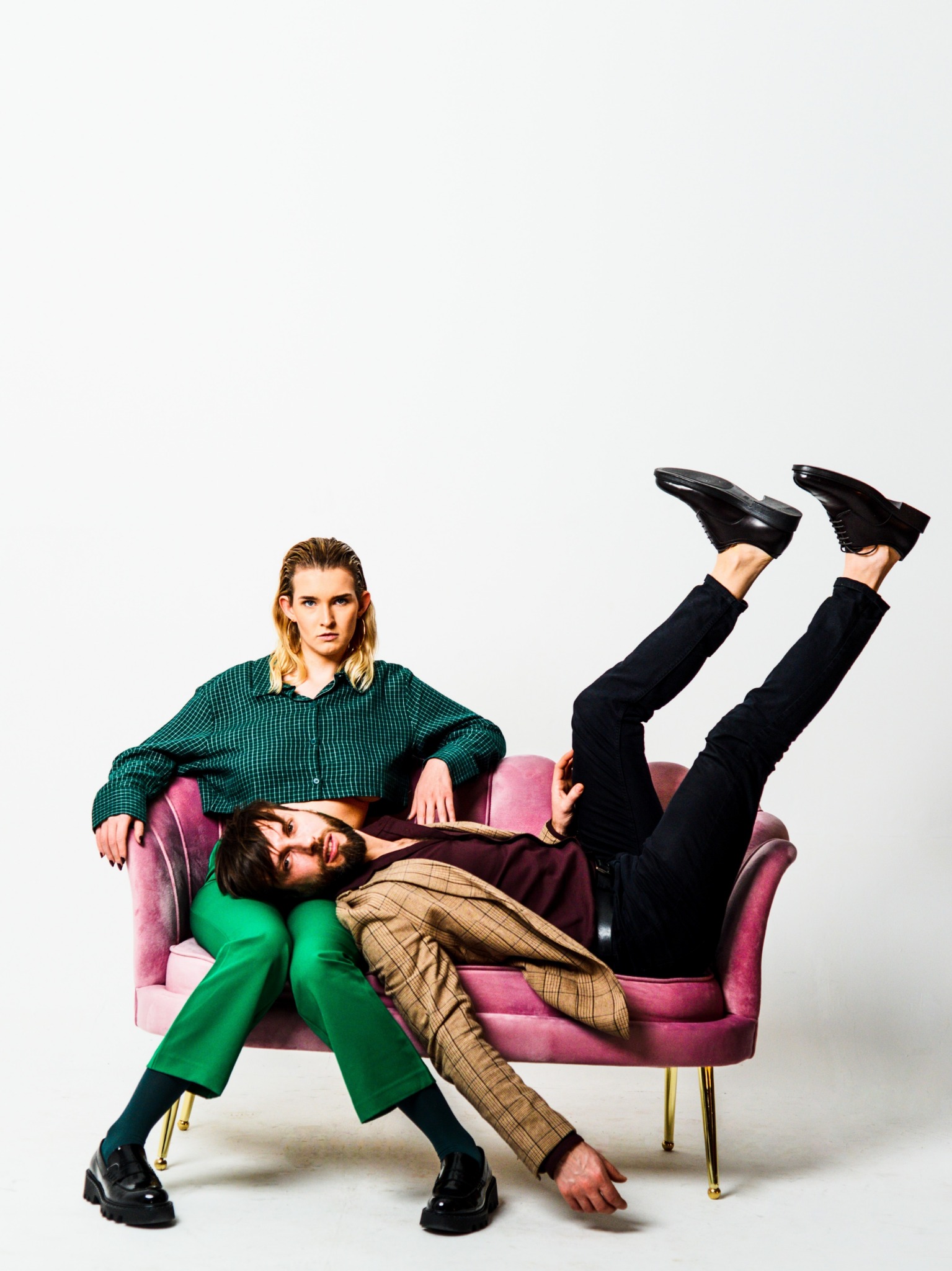Portret dwojga modeli w studyjnej sesji: kobieta w zielonej koszuli i spodniach siedzi na różowym fotelu, podczas gdy mężczyzna w brązowej marynarce leży na jej kolanach, unosząc nogi w czarnych...