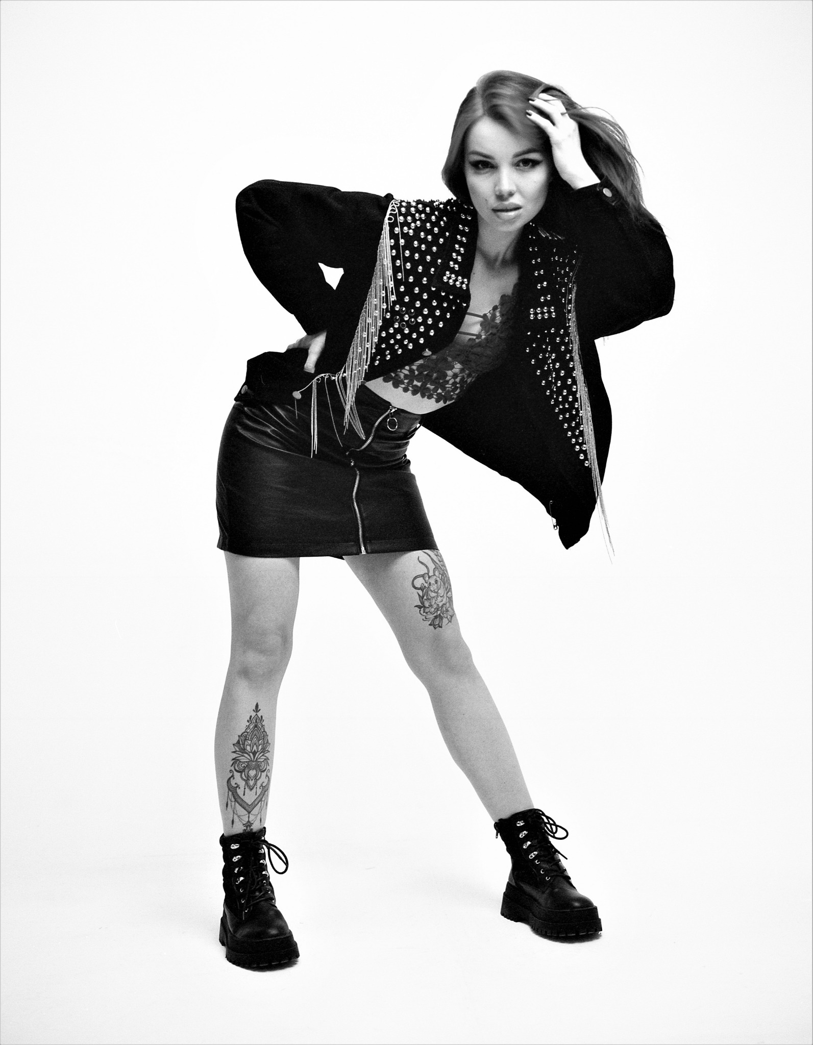 Czarno-białe zdjęcie kobiety w skórzanej mini spódniczce, koronkowej bieliźnie i zdobionej ćwiekami kurtce, pozującej na białym tle z tatuażami na nogach i w ciężkich butach.