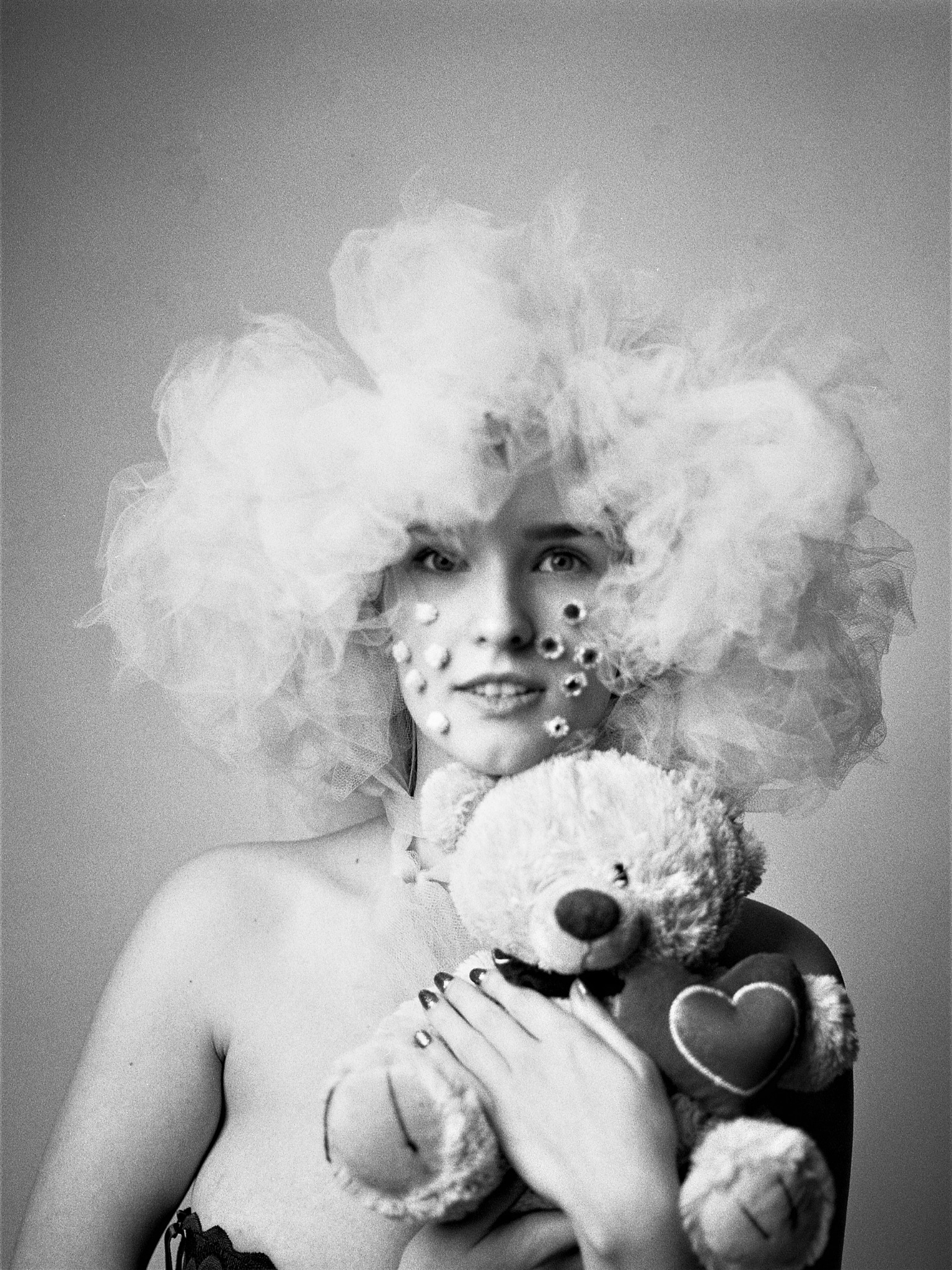 Czarno-biała fotografia studyjna z Zduńskiej Woli: kobieta z tiulową fryzurą, kwiatami na twarzy i pluszowym misiem z sercem.