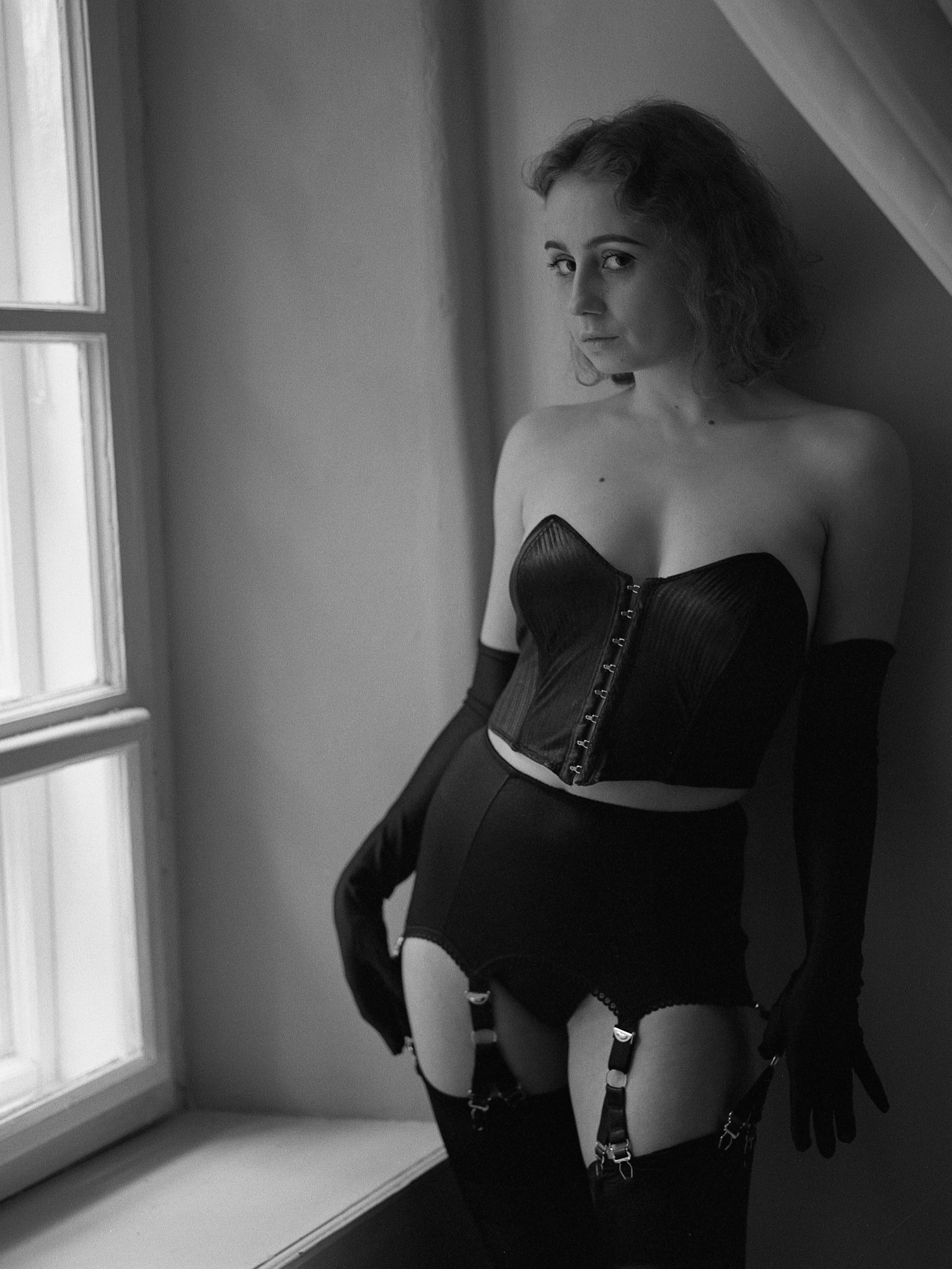Czarno-biała fotografia kobiety w bieliźnie (gorset, pas do pończoch, rękawiczki) stojącej przy oknie, z delikatnym uśmiechem i spojrzeniem w obiektyw, w pomieszczeniu z ukośnym sufitem.
