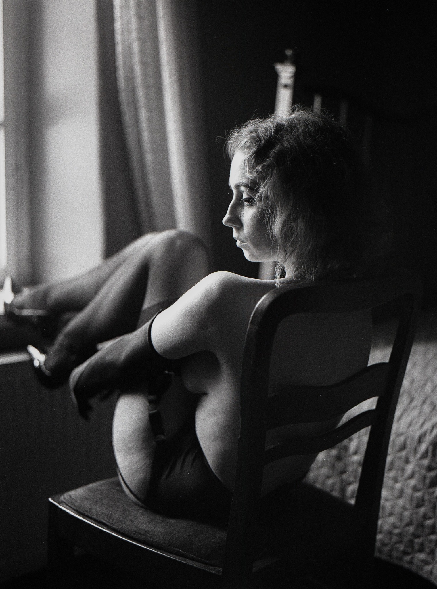 Czarno-białe zdjęcie kobiety siedzącej na krześle w pończochach i podwiązkach, odwróconej profilem w stronę okna z delikatnym światłem.