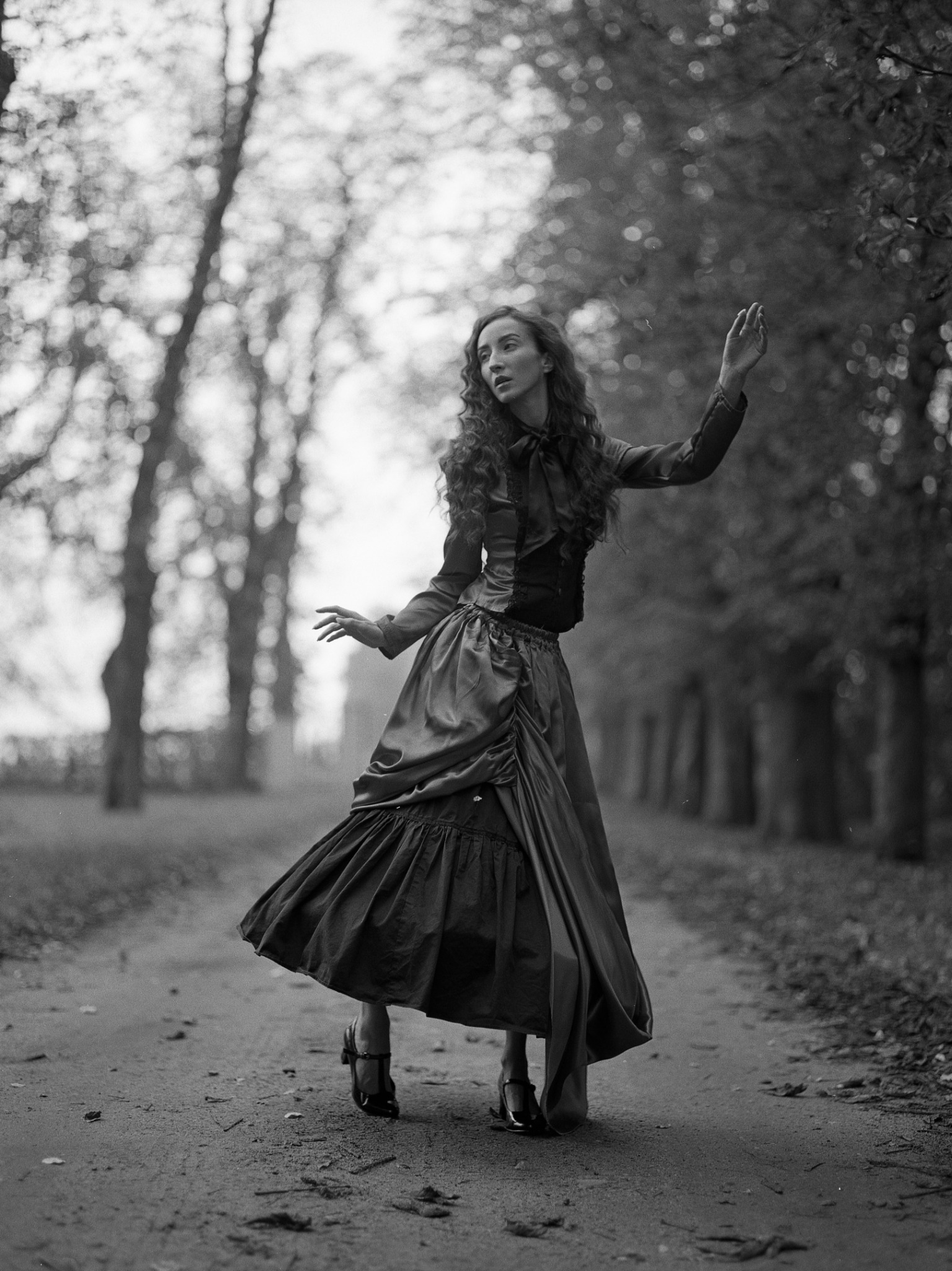 Czarno-białe zdjęcie kobiety w długiej sukni i butach na obcasie, stojącej na ścieżce w parku z podniesionymi rękami, z drzewami w tle.