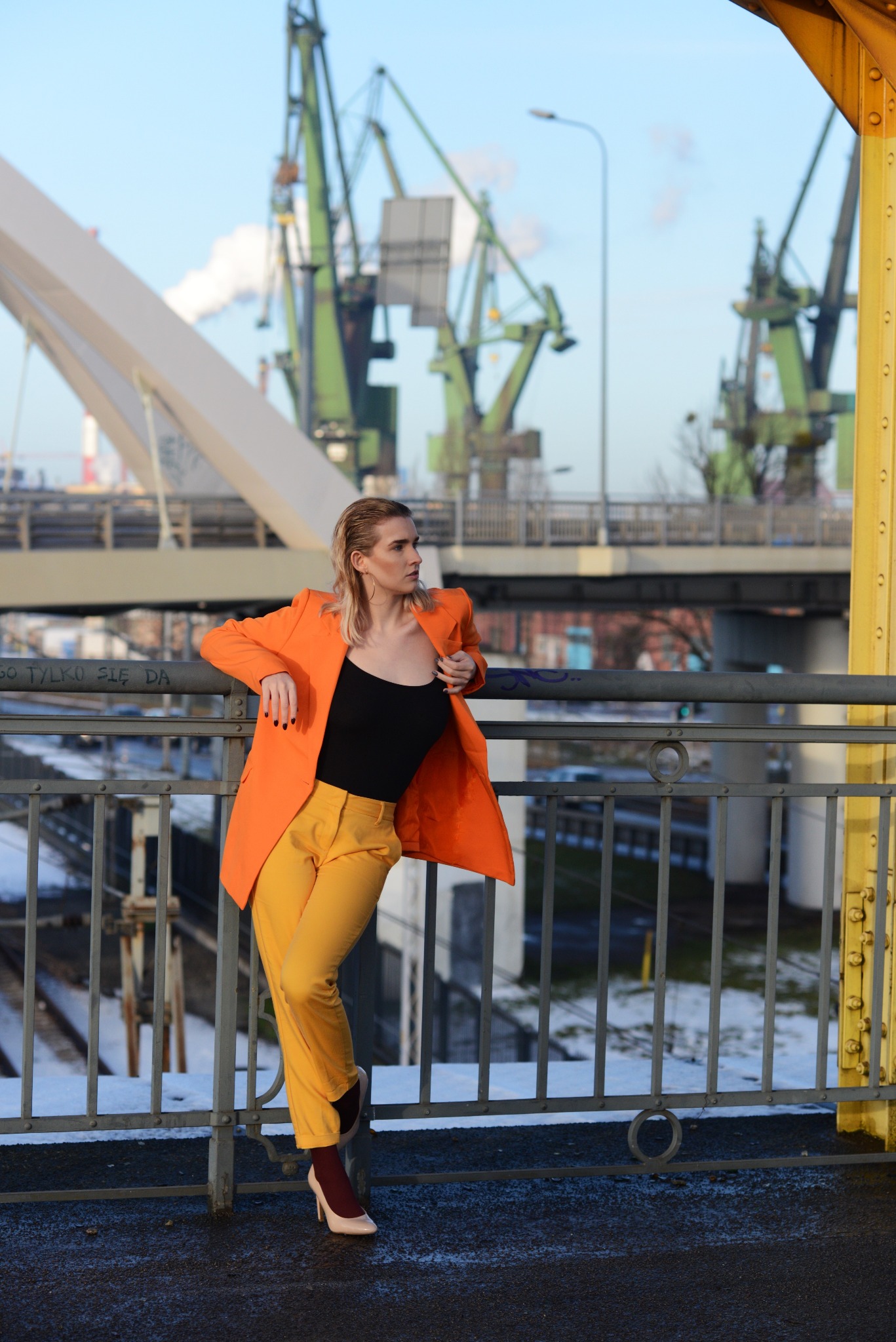 Kobieta w pomarańczowej marynarce i żółtych spodniach opiera się o barierkę mostu na tle dźwigów portowych.
