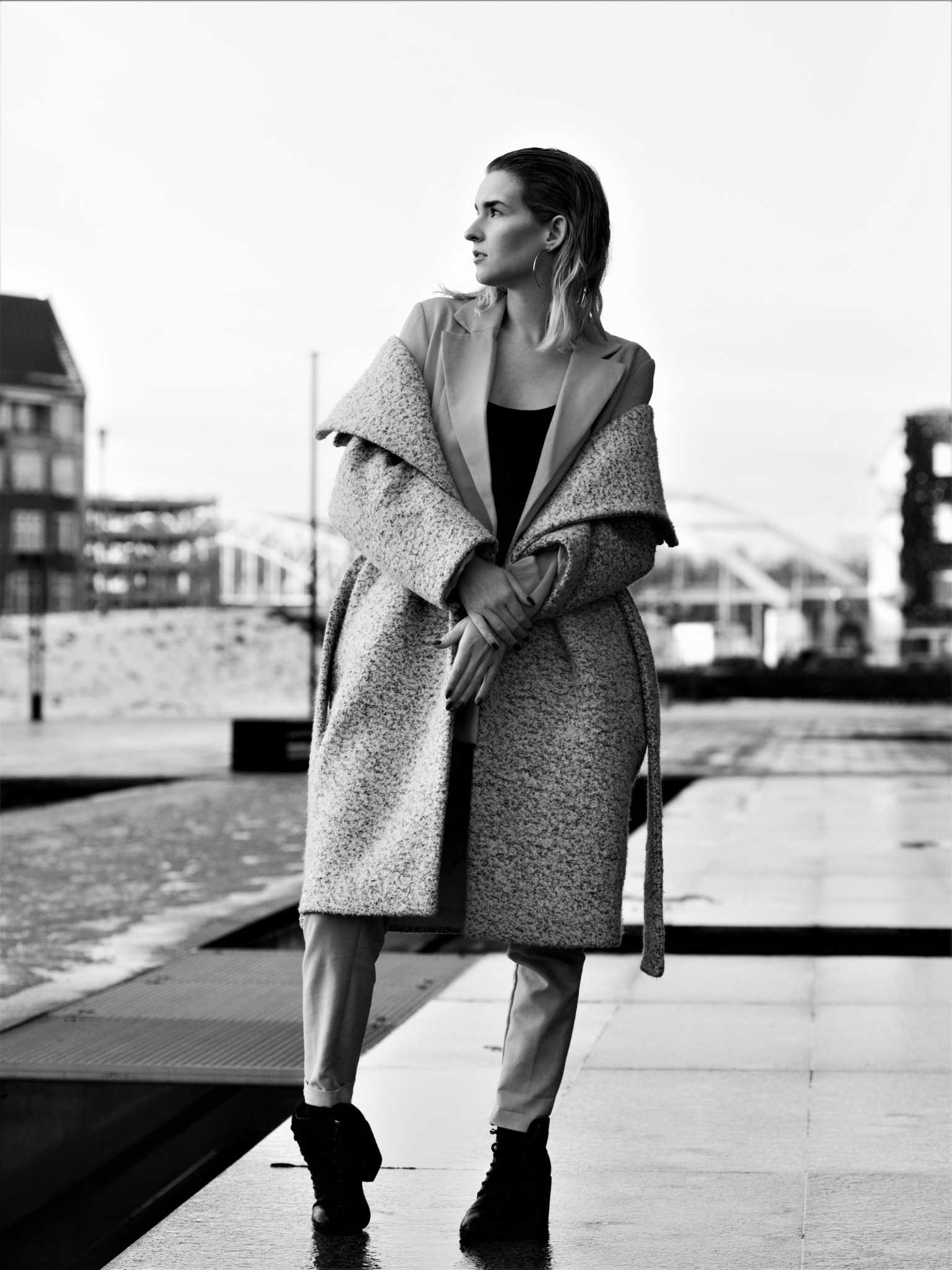 Czarno-białe zdjęcie kobiety w płaszczu z szerokimi klapami, stojącej na tle miejskiego krajobrazu z mostem w oddali, oświetlonej naturalnym światłem.