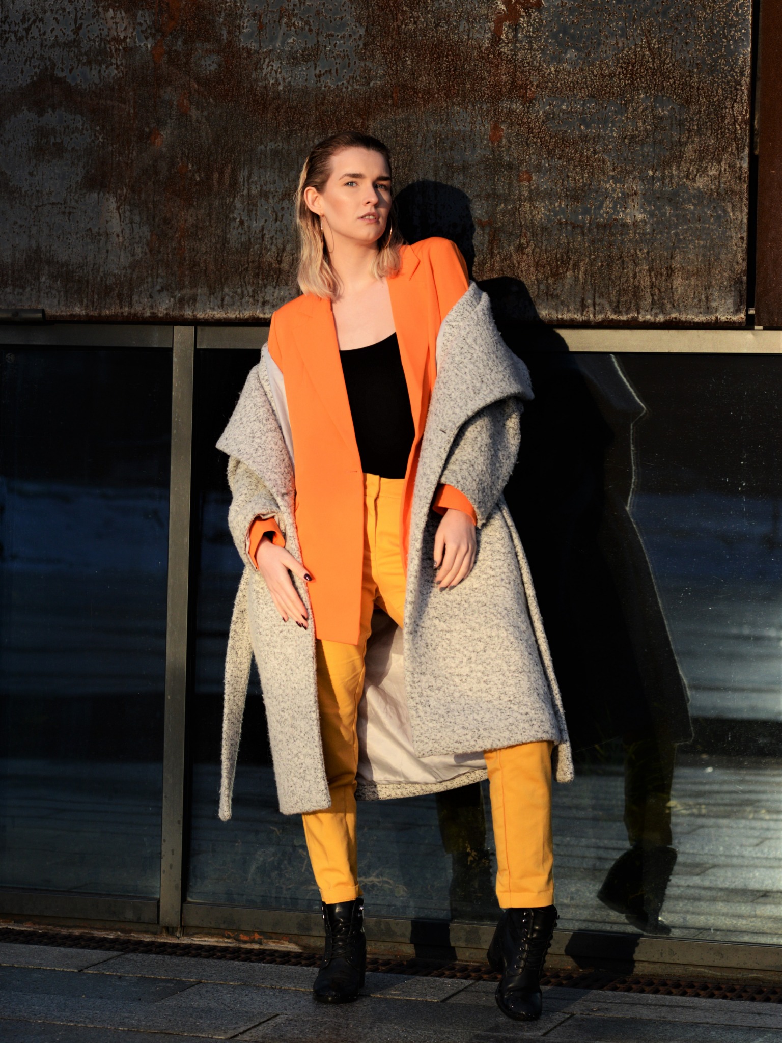 Kobieta w pomarańczowym żakiecie, żółtych spodniach i szarym płaszczu pozuje na tle metalicznej ściany z odbiciem w szybie.