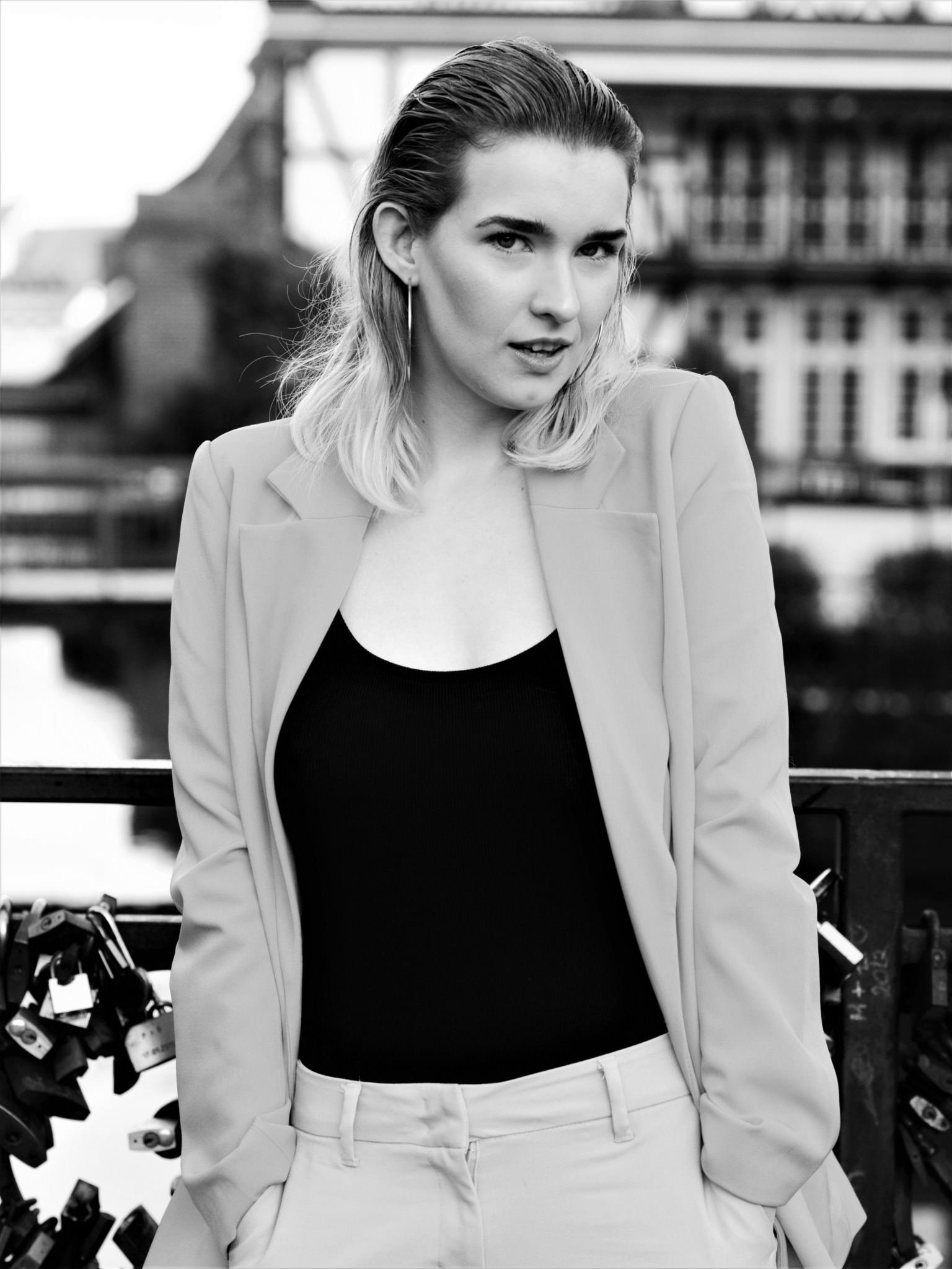 Czarno-białe portretowe ujęcie kobiety w marynarce i topie na tle miejskiego krajobrazu z mostem i kłódkami.