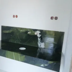 Montaż ciemnozielonego blatu z kamienia z otworem na umywalkę, widoczne puszki elektryczne w ścianie, surowy stan wykończenia.