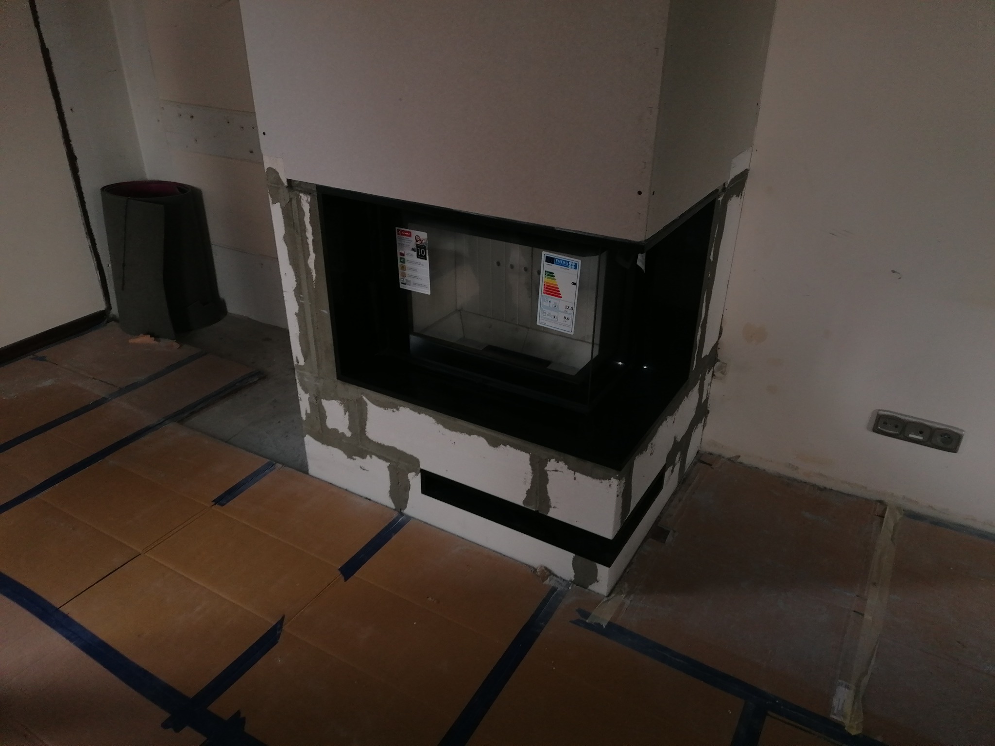 Wnętrze w trakcie wykańczania z widocznym kominkiem w obudowie z płyt gipsowych, zabezpieczone kartonem na podłodze.