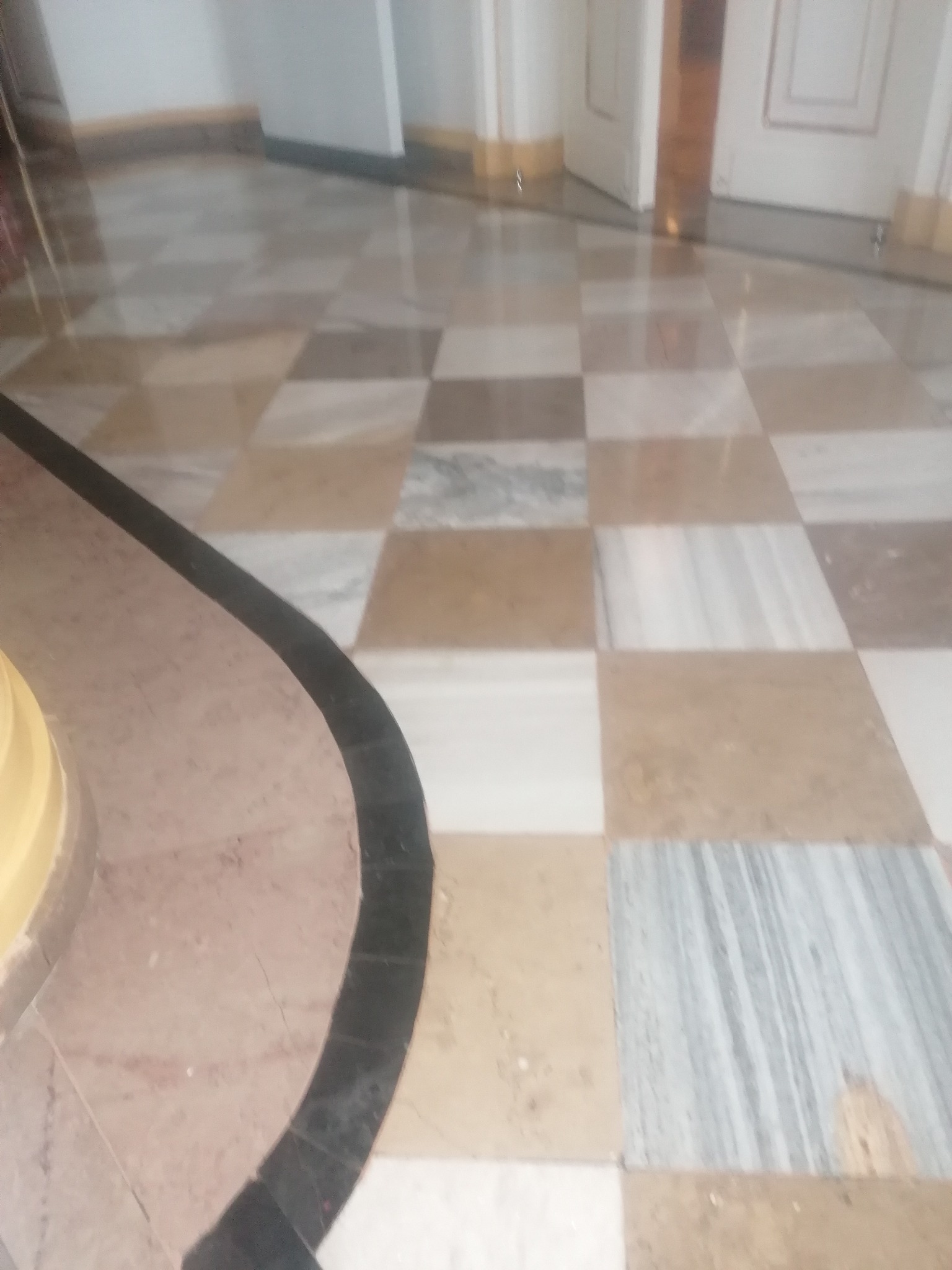 Szachownica z polerowanych, beżowo-szarych płyt kamiennych na podłodze, z widocznym fragmentem zakrzywionej krawędzi z ciemną obwódką i białymi drzwiami w tle.