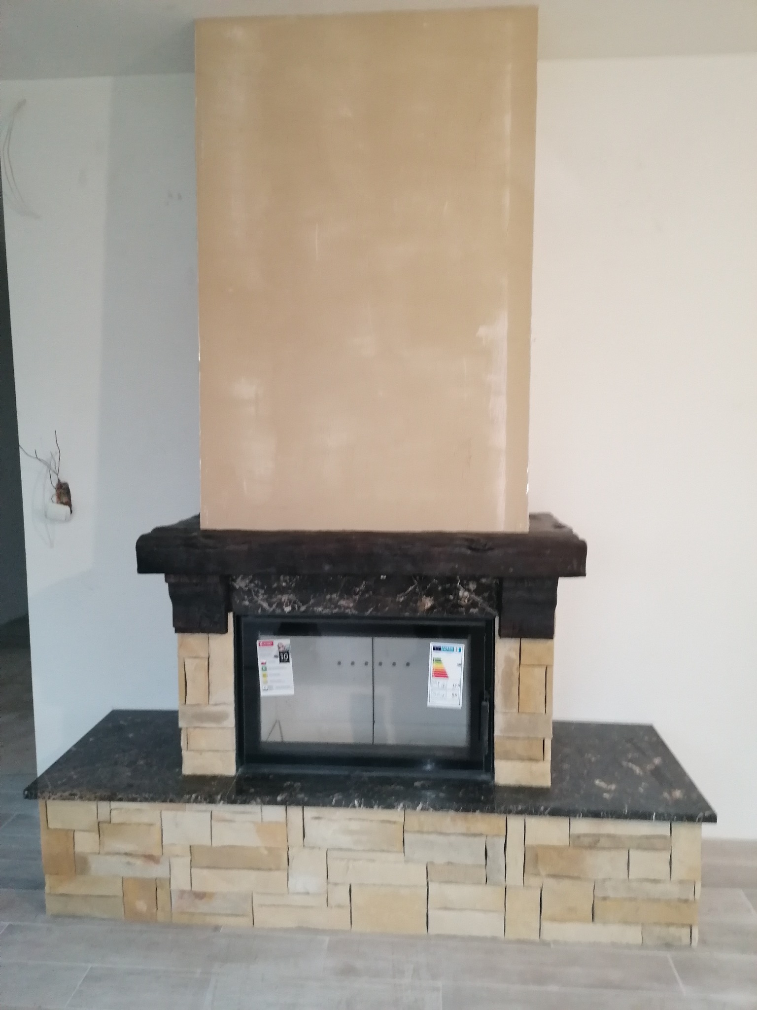 Wnętrze z kominkiem obudowanym kamieniem naturalnym i elementami z ciemnego drewna, ściana nad kominkiem przygotowana do wykończenia, widoczne wystające kable.