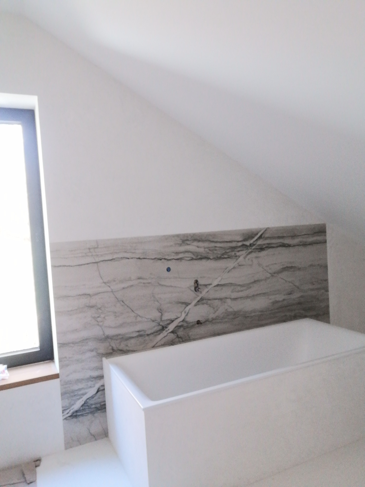 Wanna w łazience w Warszawie z obudową z płyty imitującej marmur, zamontowana pod skosem dachu, widoczne okno.