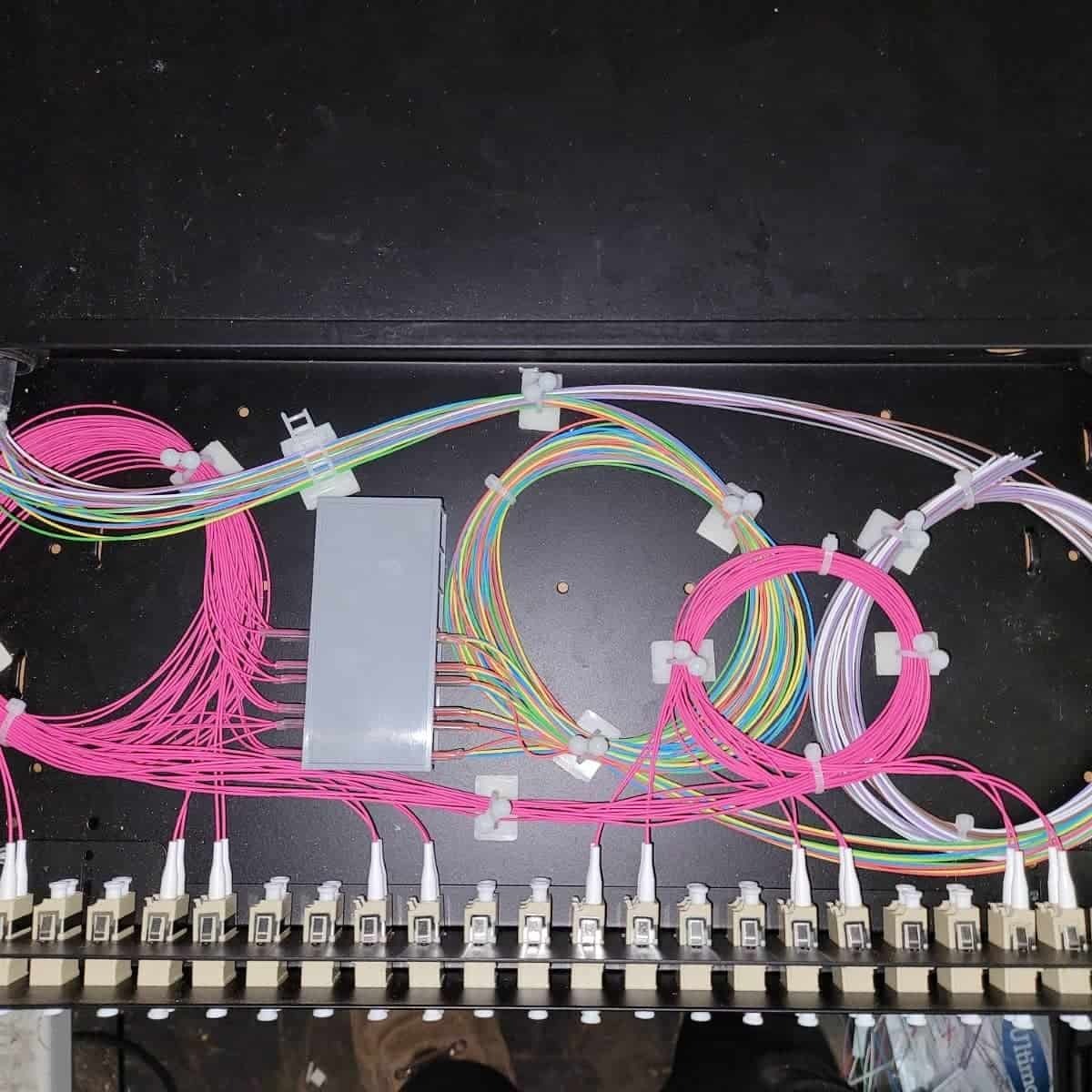Panel światłowodowy z uporządkowanymi kablami w kolorach tęczy i różu, zabezpieczonymi białymi uchwytami, widok z góry.