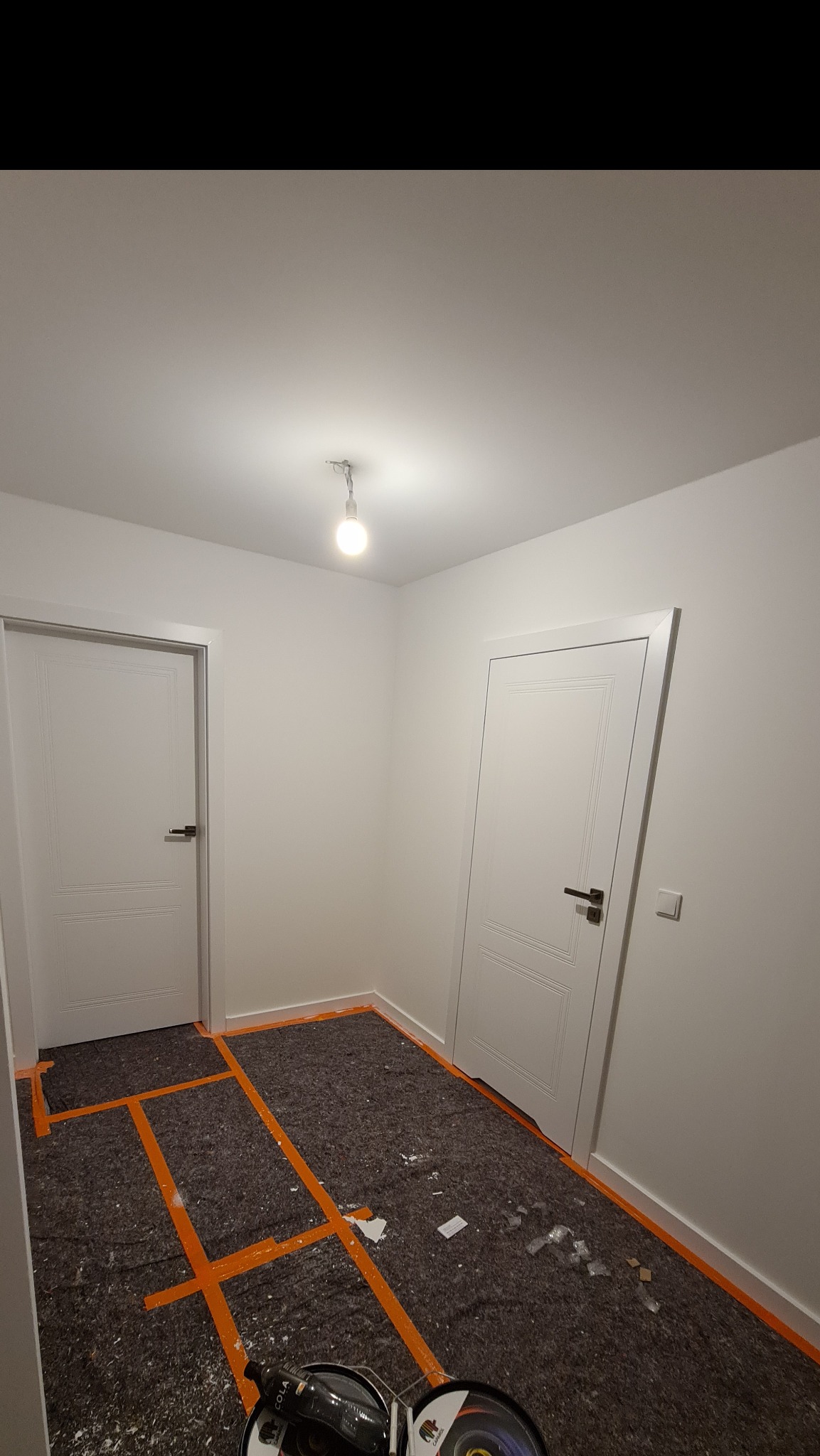 Przygotowanie podłoża w pomieszczeniu z dwiema białymi drzwiami, zabezpieczone folią i taśmą malarską, widoczne narzędzia i resztki materiałów budowlanych.