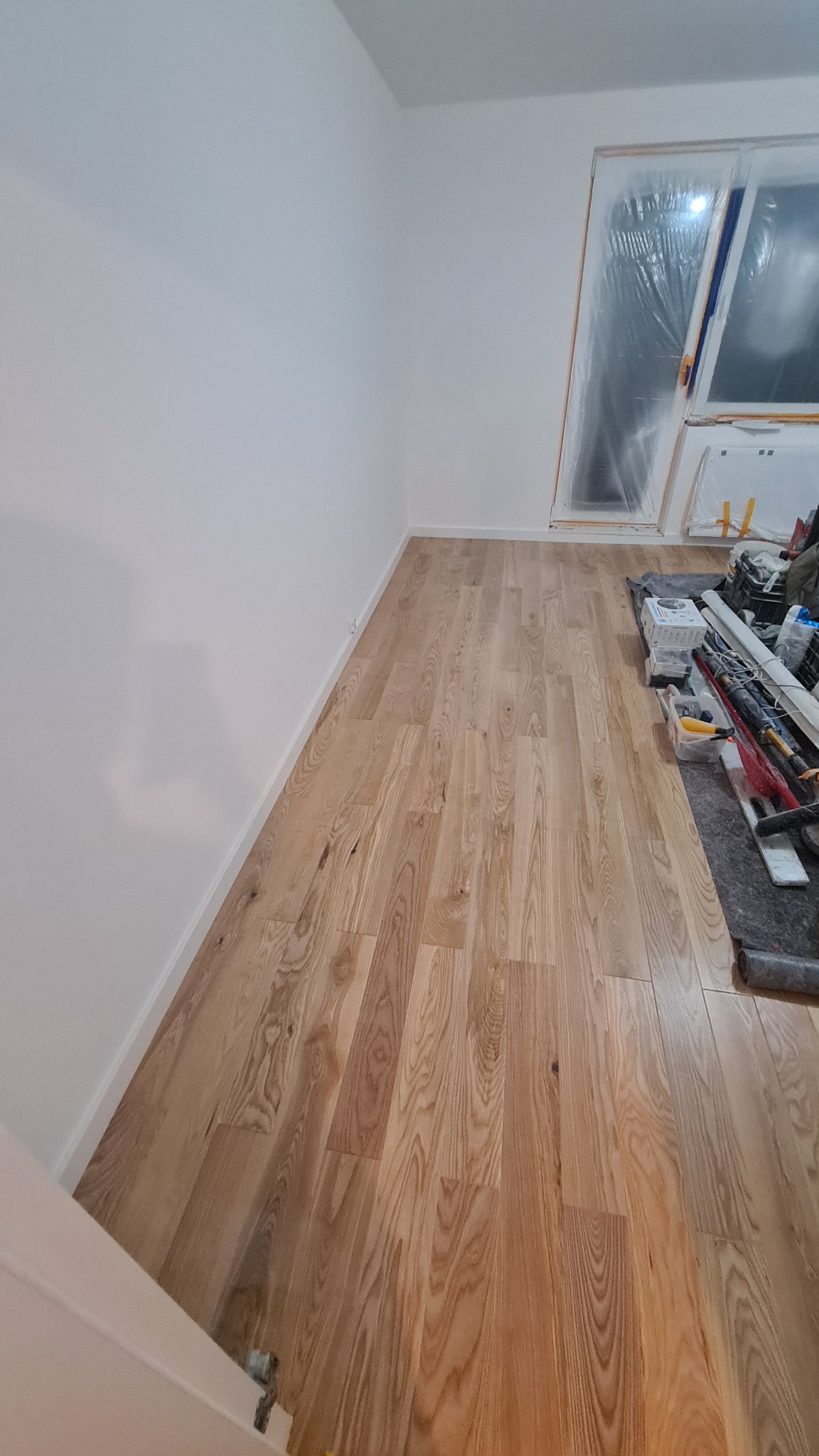 Nowo położona, jasna, drewniana podłoga w pokoju z pomalowanymi na biało ścianami i oknem zabezpieczonym folią malarską, widoczne narzędzia i materiały budowlane.