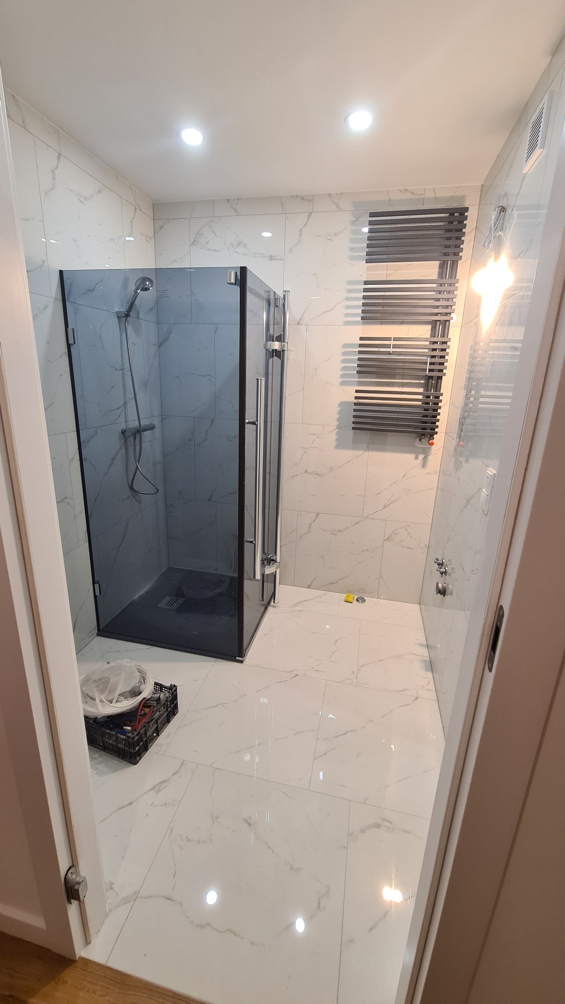 Nowoczesna łazienka z kabiną prysznicową o ciemnych szybach, grzejnikiem drabinkowym i białymi płytkami imitującymi marmur, widoczne narzędzia w skrzynce na podłodze sugerują trwające prace.