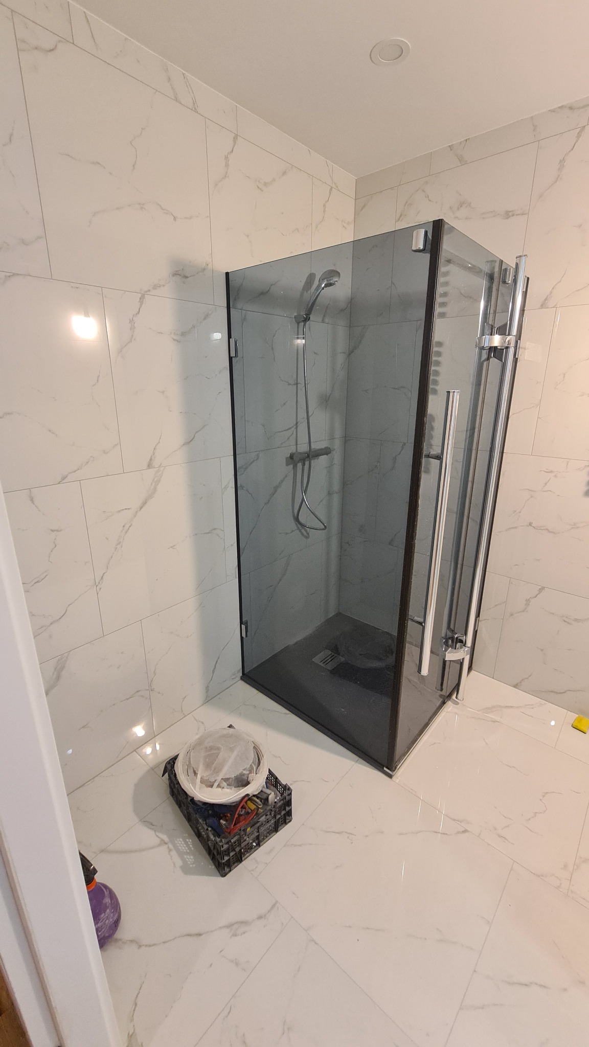 Szklana kabina prysznicowa z czarnymi ramami w łazience wyłożonej marmurowymi płytkami, widoczny koszyk z narzędziami i akcesoriami glazurniczymi obok kabiny.