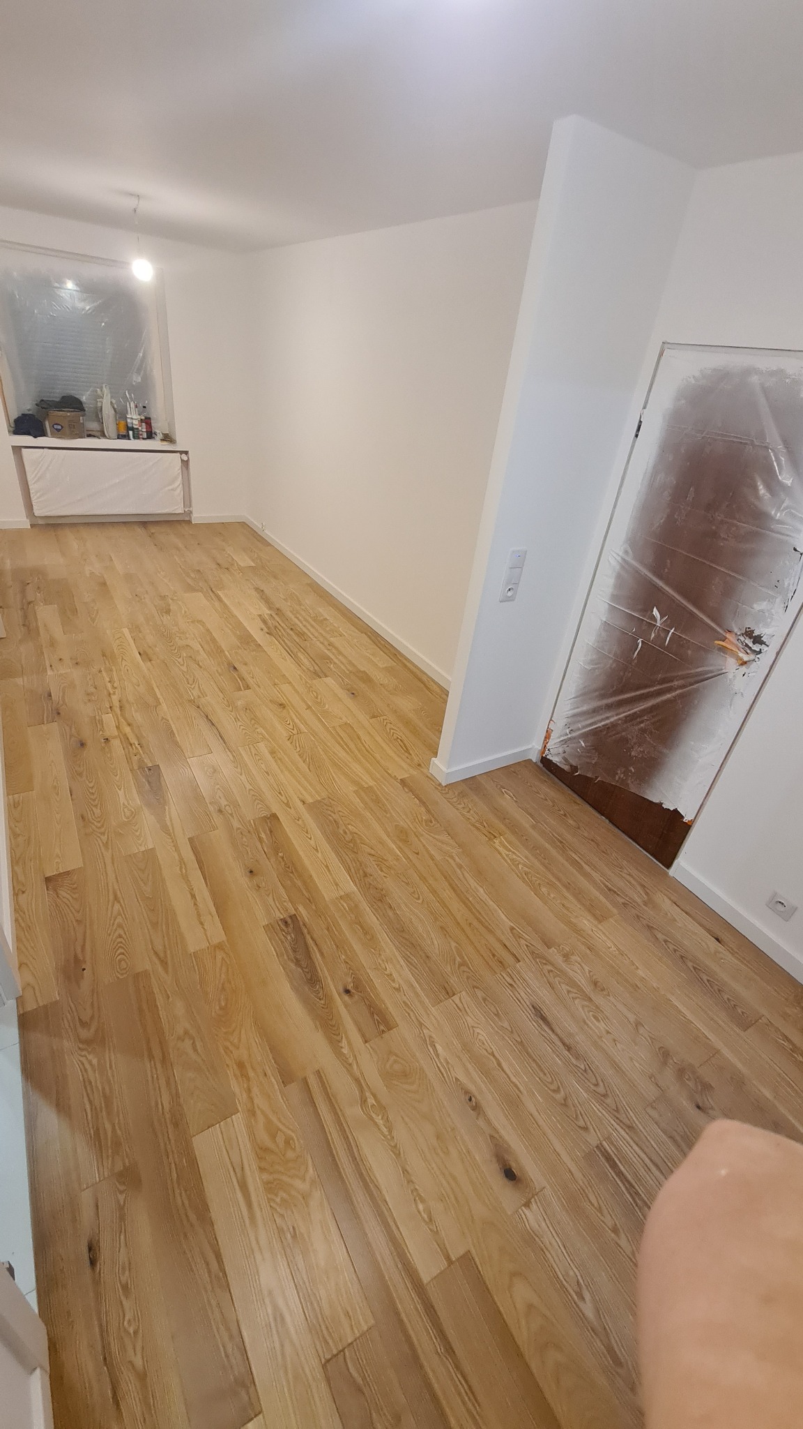 Pokój po remoncie z nową drewnianą podłogą, okno i drzwi zabezpieczone folią malarską, ściany pomalowane na biało.