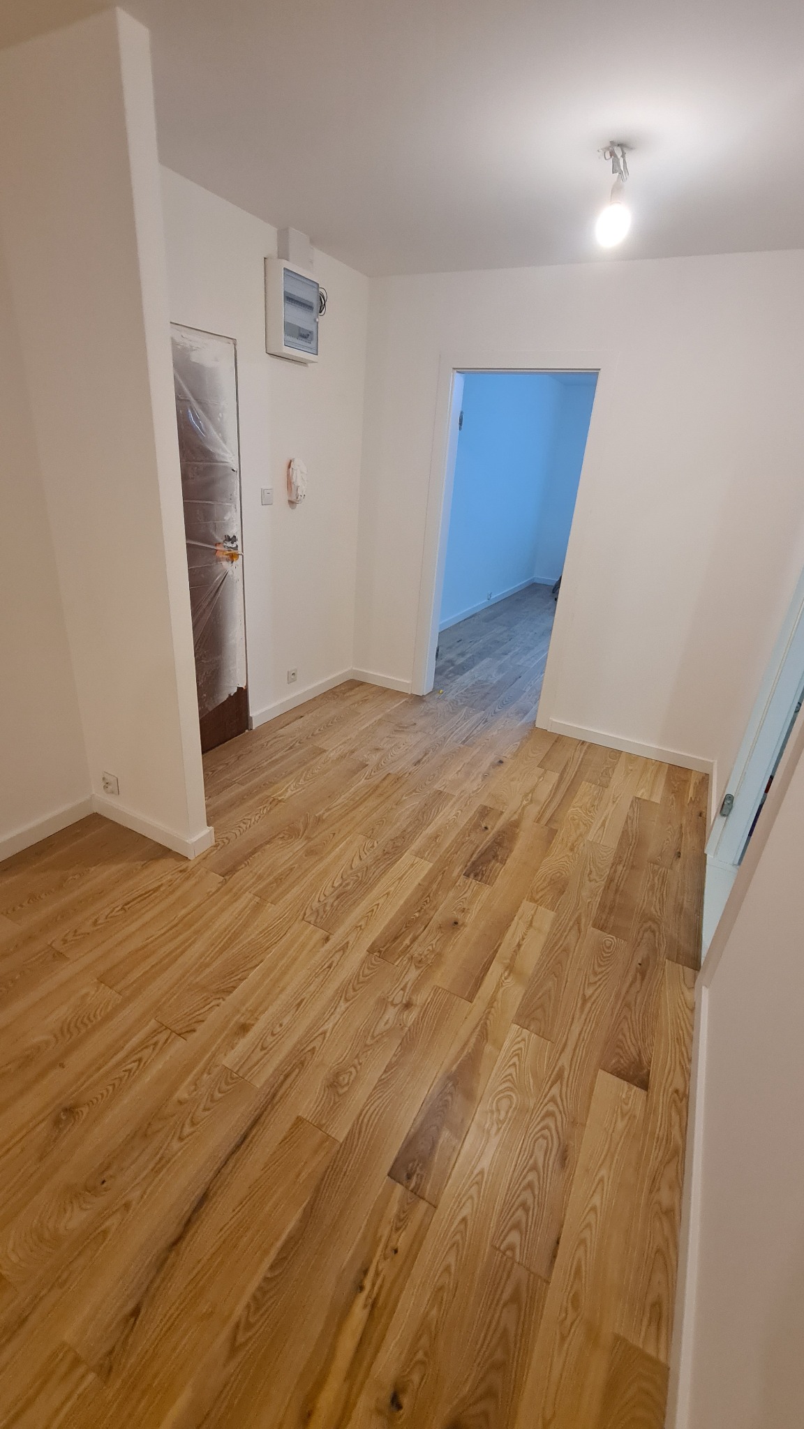 Jasne, drewniane panele podłogowe ułożone w nowym mieszkaniu, widok na otwarte drzwi do kolejnego pomieszczenia z identyczną podłogą.