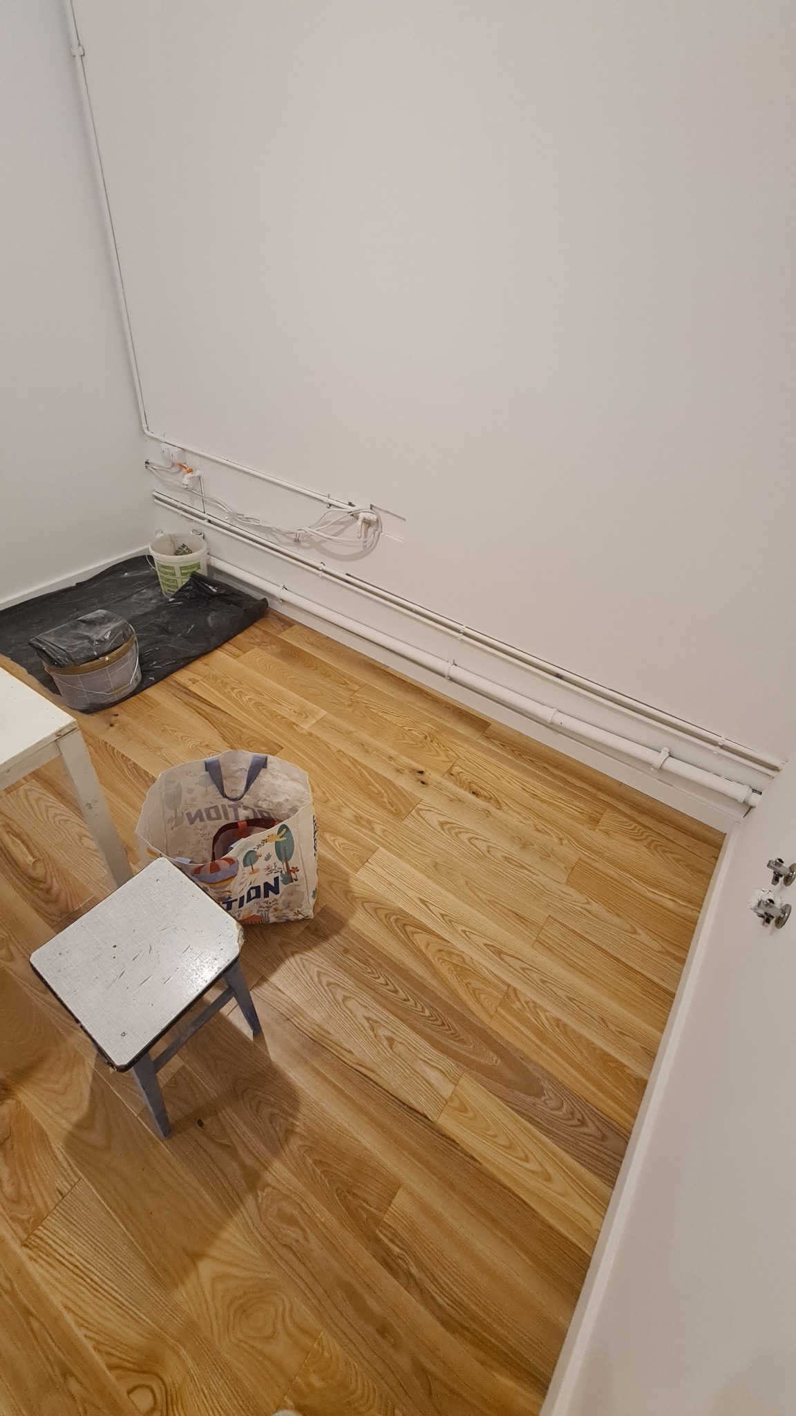 Przygotowania do prac wykończeniowych w pomieszczeniu: wiadro z farbą, folia ochronna, torba z narzędziami i drewniana podłoga z wyraźnym usłojeniem, widoczne rury instalacyjne pod sufitem.