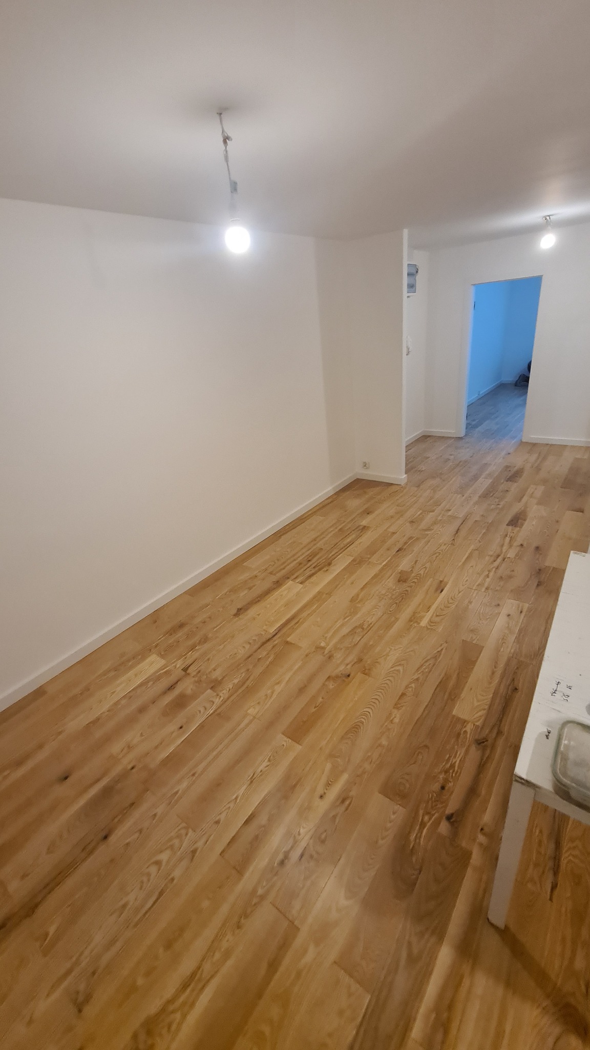 Puste pomieszczenie po remoncie z nową drewnianą podłogą i świeżo pomalowanymi białymi ścianami, widoczna żarówka na kablu zwisająca z sufitu, w tle otwarte drzwi do kolejnego pomieszczenia.