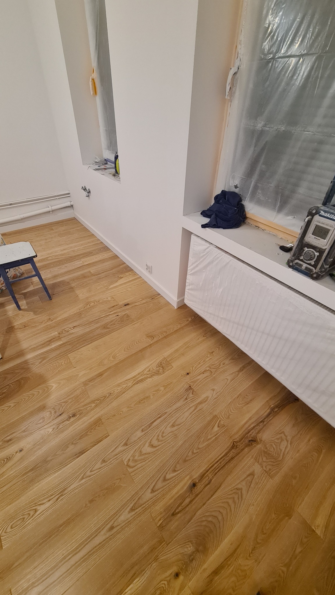 Parkiet ułożony w jodełkę, zabezpieczony grzejnik folią malarską, okno zakryte folią, widoczny radiobudzik Makita i niebieska bluza.