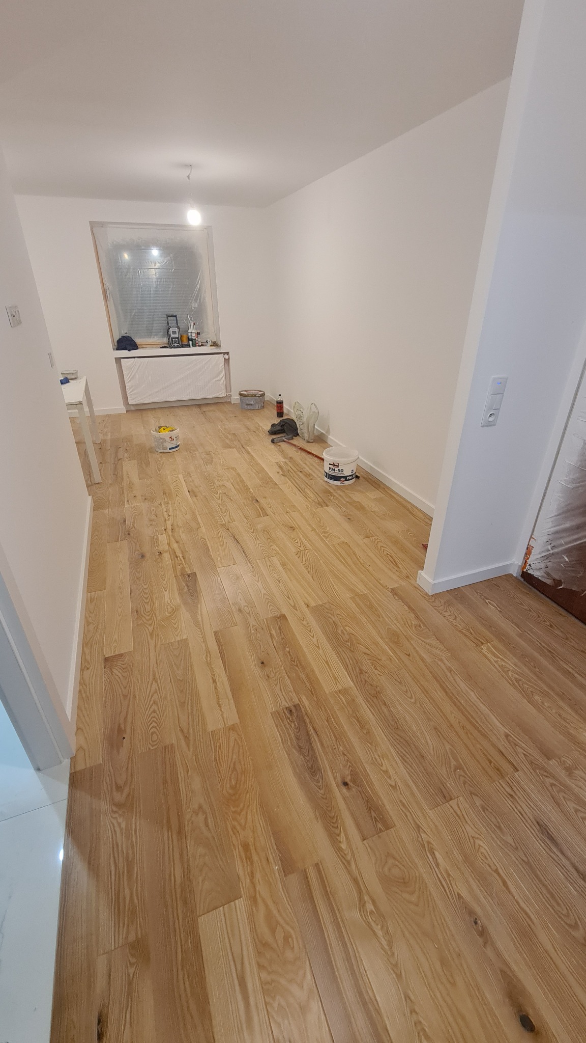 Pokój po malowaniu z drewnianą podłogą, okno i grzejnik zabezpieczone folią malarską, widoczne wiaderka z farbą i akcesoria malarskie.