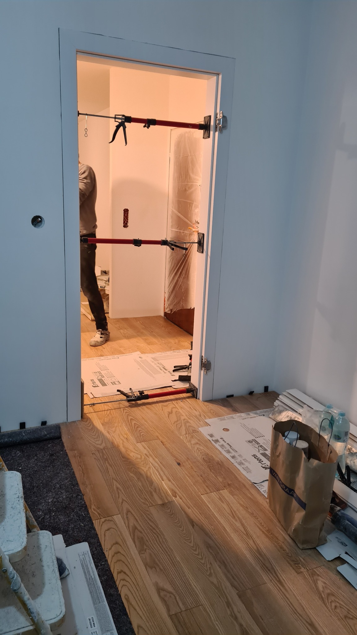Montaż ościeżnicy drzwiowej z użyciem rozpórek teleskopowych, widoczna osoba w tle, podłoga zabezpieczona kartonem, detale wykończeniowe w trakcie prac.