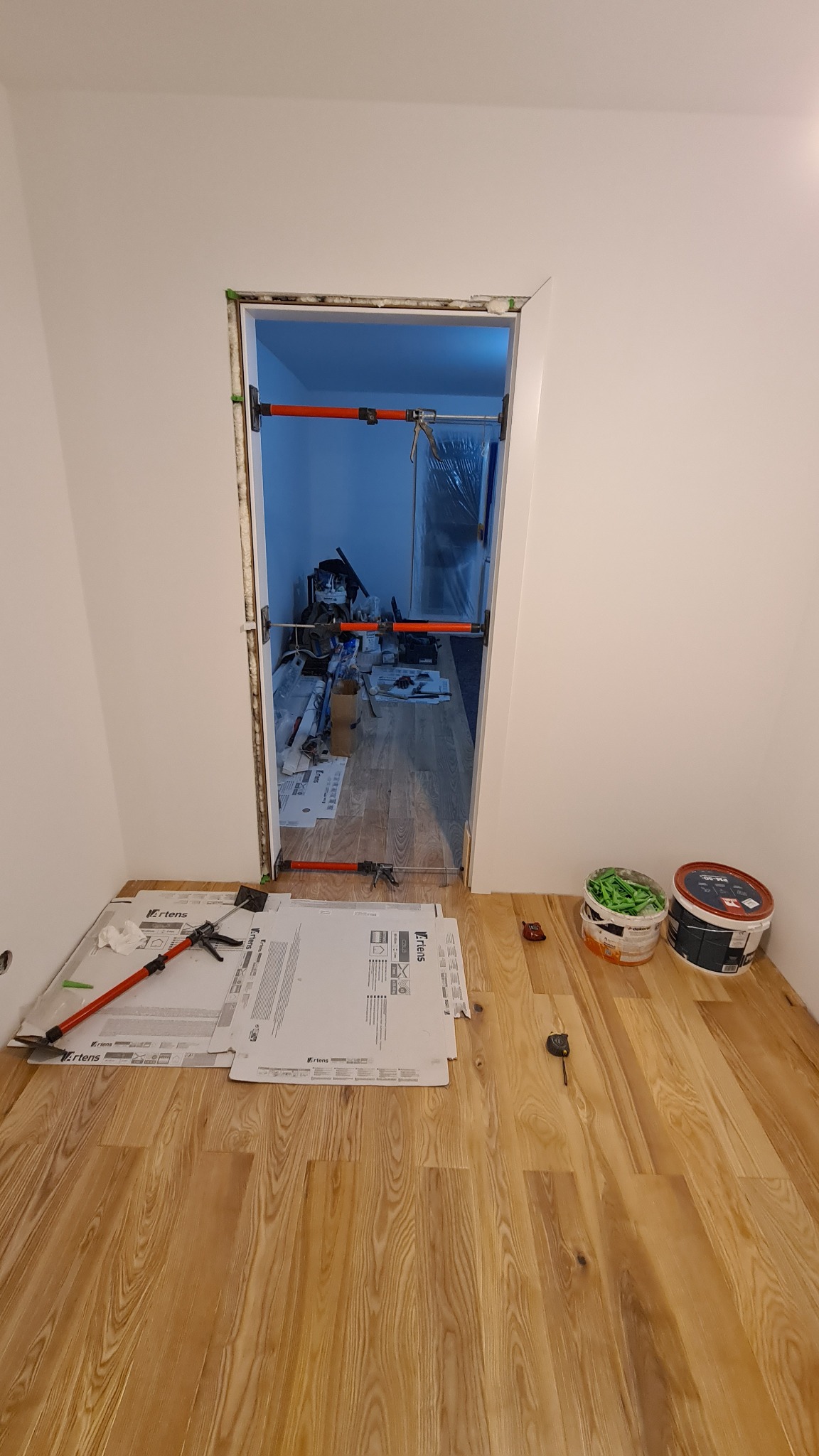 Przygotowanie do montażu ościeżnicy drzwiowej: widoczne rozpórki, puszki z akrylem i kartony ochronne na parkiecie.