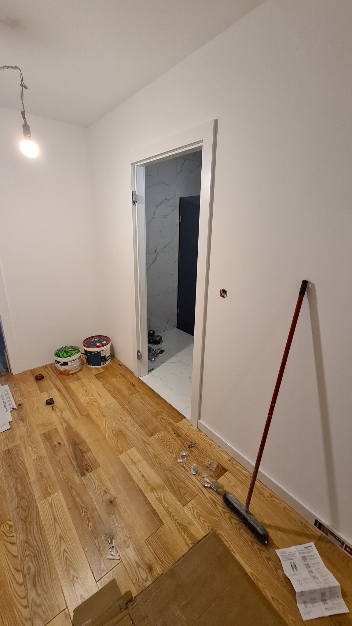 Wnętrze w trakcie prac wykończeniowych: drewniana podłoga, białe ściany z widocznym otworem drzwiowym do wykończonej łazienki z marmurowymi płytkami, puszki farb, szczotka i rozłożona instrukcja...