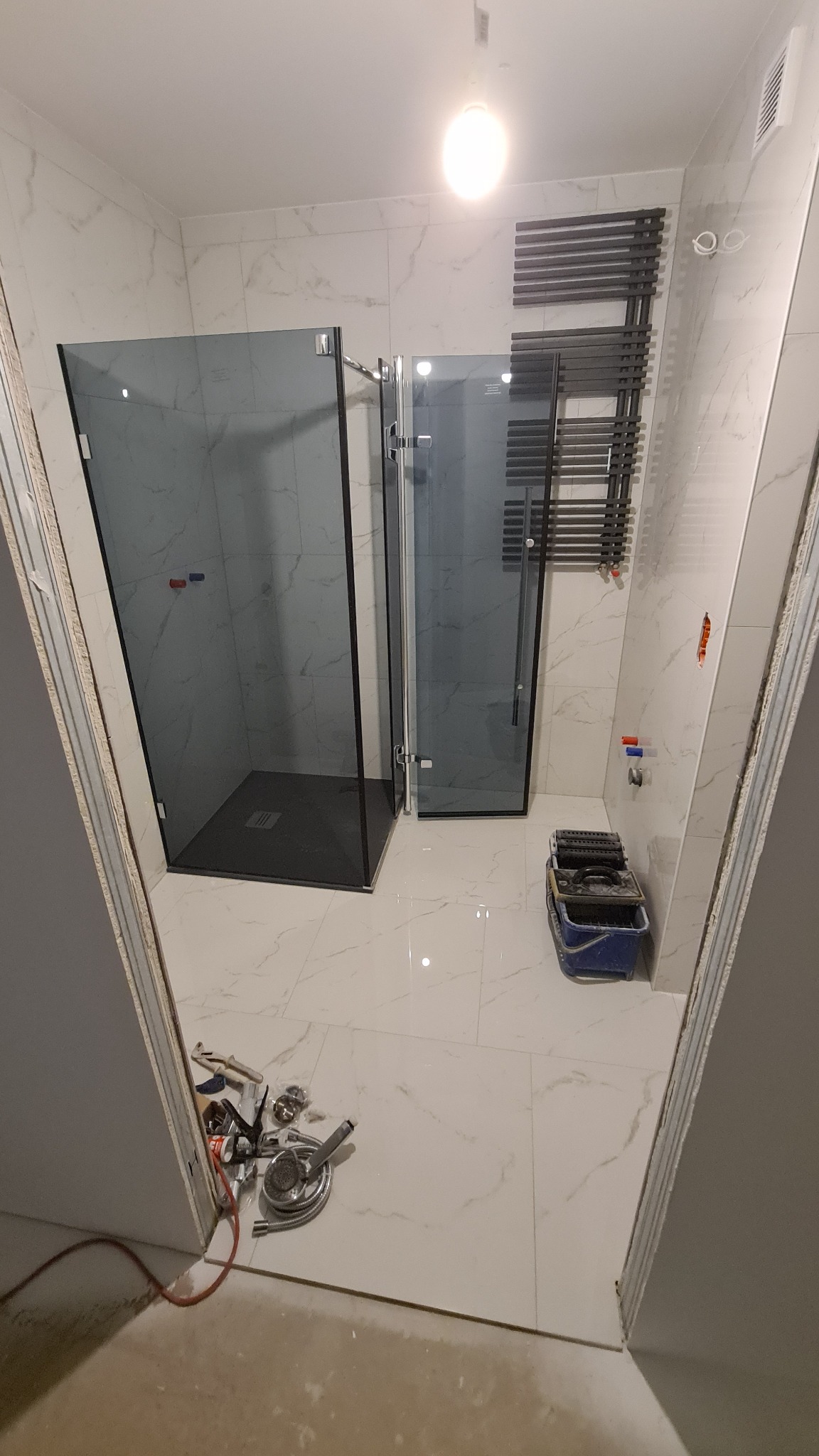 Nowoczesna kabina prysznicowa z czarnym brodzikiem i szklanymi drzwiami na tle marmurowych płytek, widoczne narzędzia glazurnicze i grzejnik drabinkowy.