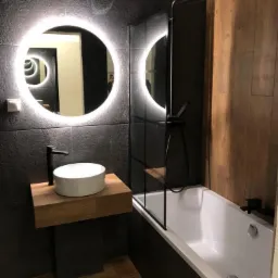 Nowoczesna łazienka z okrągłym lustrem LED, umywalką nablatową na drewnianym blacie i wanną z czarną kabiną prysznicową.