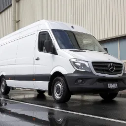 Biały, dostawczy Mercedes-Benz Sprinter na mokrej nawierzchni, odbijający się w kałużach. Widok z przodu, na tle budynku przemysłowego z blachy falistej.