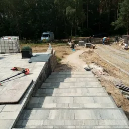 Widok z góry na częściowo ukończone schody z kostki brukowej prowadzące do ogrodu; w tle las, betoniarka i pracownicy.