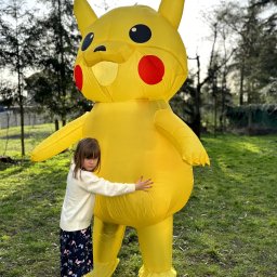 KIDS CLUB Szymon Wyrwas - Dziewczynka w jasnym swetrze i wzorzystej spódnicy przytula dmuchany kostium Pikachu na trawniku w plenerze.