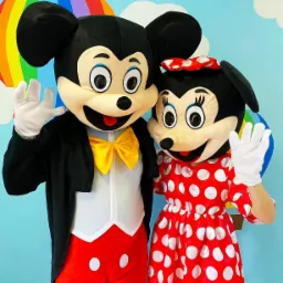 Dwie osoby w kostiumach Myszki Miki i Minnie machają rękami na tle błękitnej ściany z tęczą i chmurami.