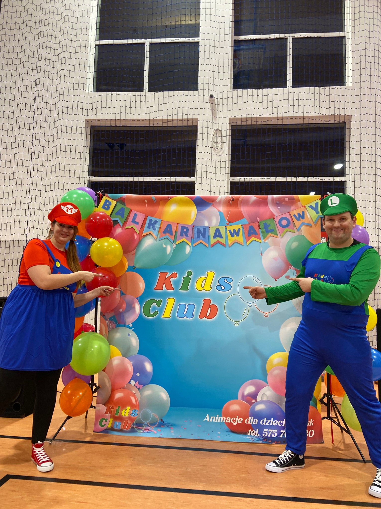 Dwie osoby przebrane za Mario i Luigi wskazują palcami na baner z napisem 'Balkarnawałowy Kids Club', otoczone kolorowymi balonami, w sali gimnastycznej z siatką ochronną w tle.
